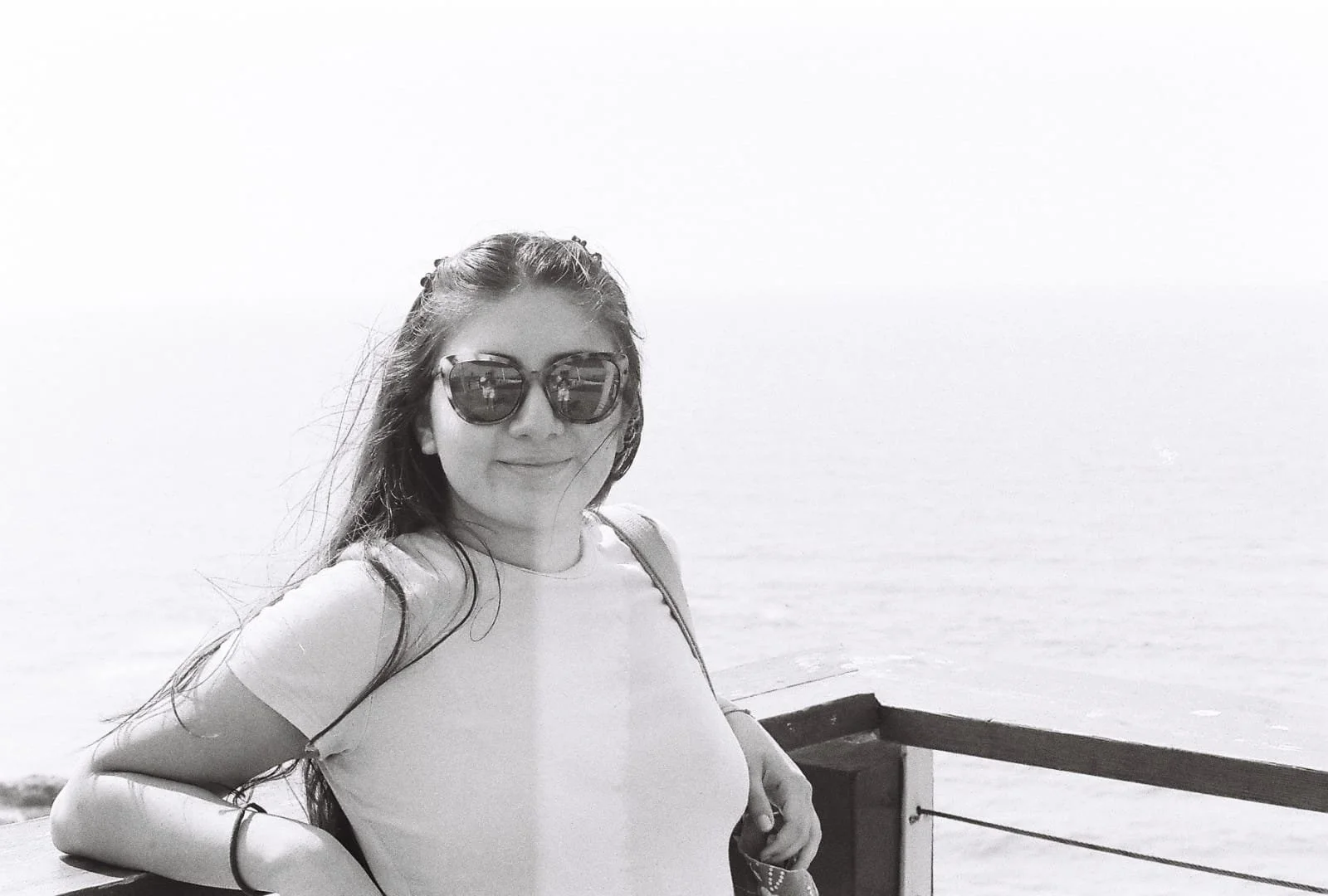 Young woman with long hair wearing sunglasses and a short-sleeve top, leaning on a wooden railing, with the ocean in the background.