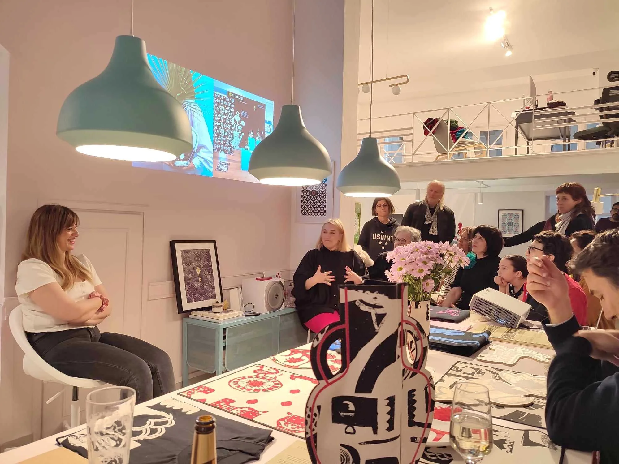 A Creative Exchange community gathering at Until Sunday Studio, a speaker sits at a long table engaging with attendees under modern pendant lights in a warm, collaborative workshop setting with a projector screen on the wall.