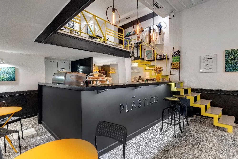 Plastico Gallery, a partner venue for the Creative Exchange hosted by Until Sunday Studio. The photo shows a sleek black bar counter with stools, warm pendant lighting, and a mezzanine loft with vibrant yellow railings.