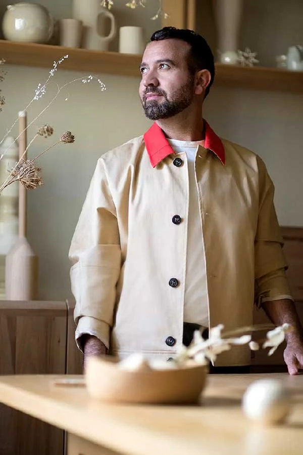 Portrait of creative consultant Antonio Aricò, a featured speaker at the Creative Exchange by Until Sunday Studio, standing in a warm, minimalist design space wearing a tan jacket with a bright red collar.