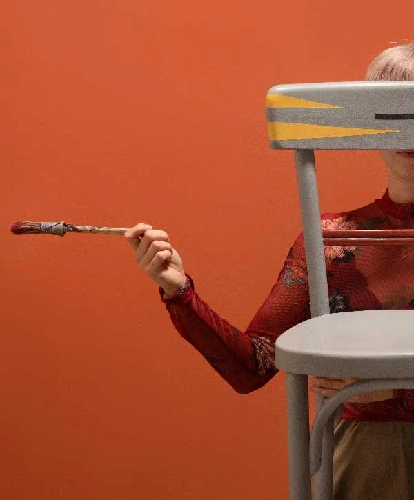 An artist holding a paintbrush behind a restored grey wooden chair from Wood Tales, featured by Until Sunday Studio's Creative Exchange.