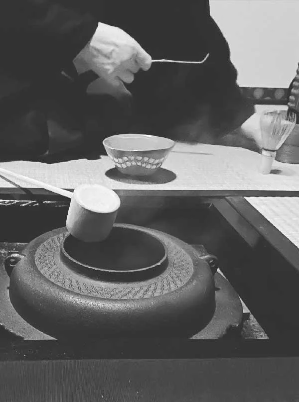 Black and white photo of a traditional Japanese tea ceremony at Kanjoan, Kyoto, featured by Until Sunday Studio's Creative Exchange. The close-up captures a tea master using a wooden ladle over a traditional iron kettle next to a ceramic matcha bowl.