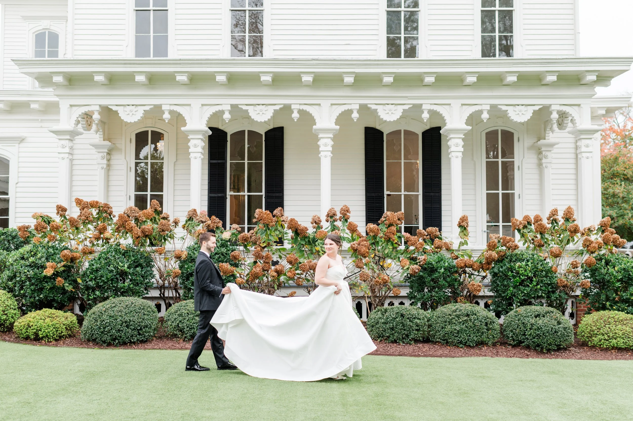 A bride and groom walk in front of the wedding venue. The groom is carrying the train of the bride's dress as he walks behind her and they look at each other.