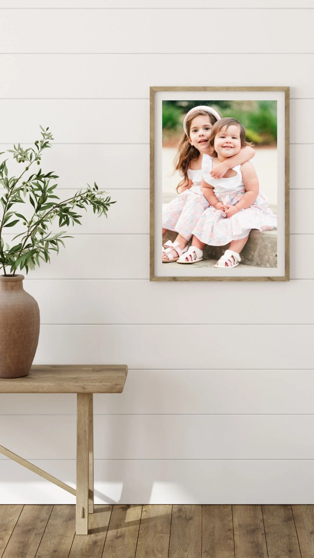 A framed photo of two young girls sitting together outdoors, smiling, with a potted plant on a small wooden table beside a white wall.