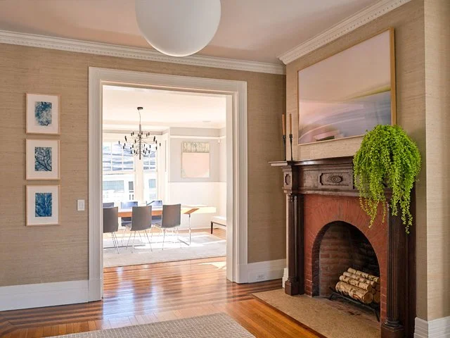 A room showcasing a fireplace to the right side with a large framed painting above. A dining room in the distance with a stacked trio of blue art on the lefthand side of the frame.