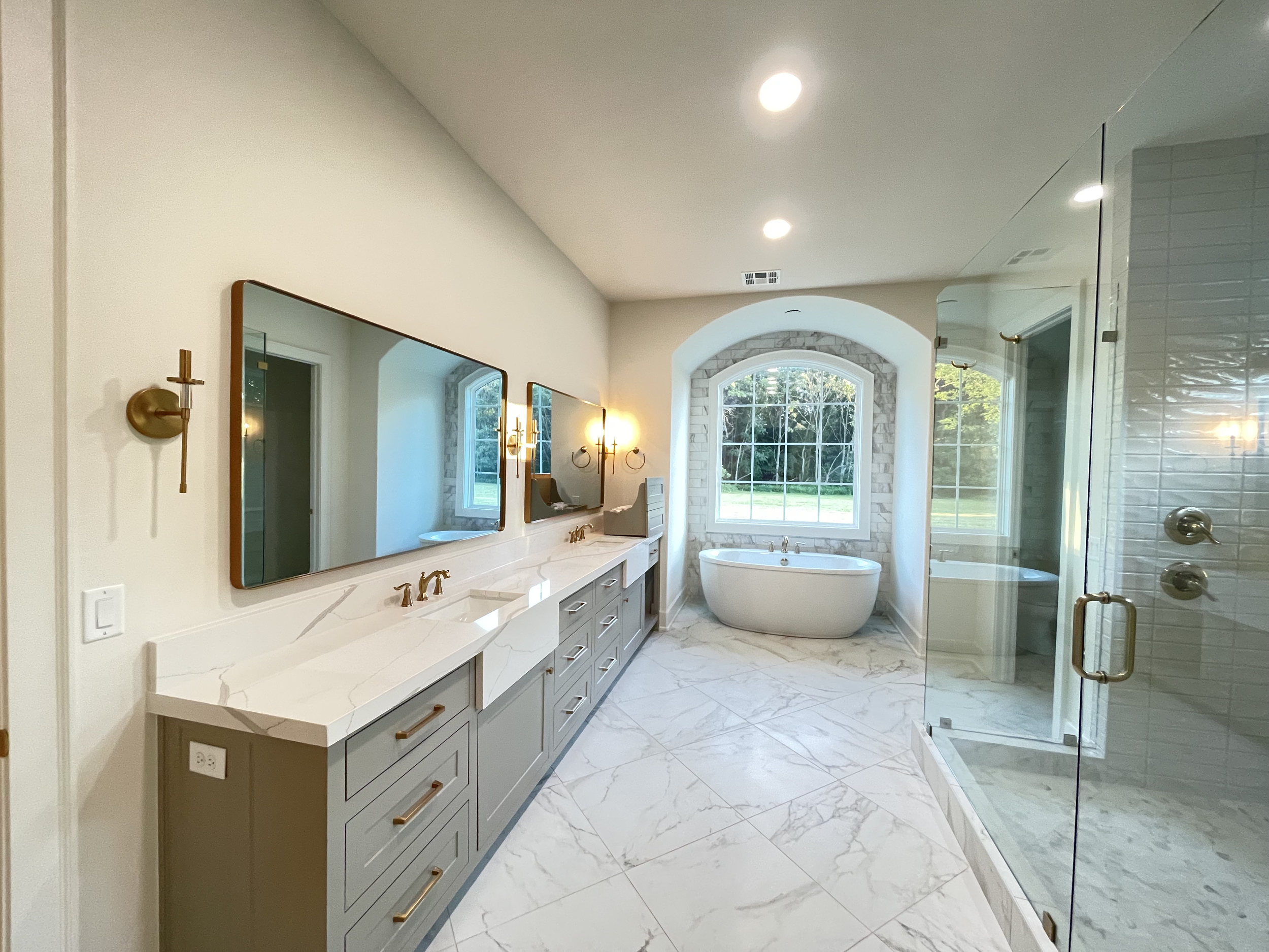 Modern bathroom with double vanity, large mirrors, brass fixtures, a free-standing bathtub near a window, and a glass-enclosed shower with brass handles.