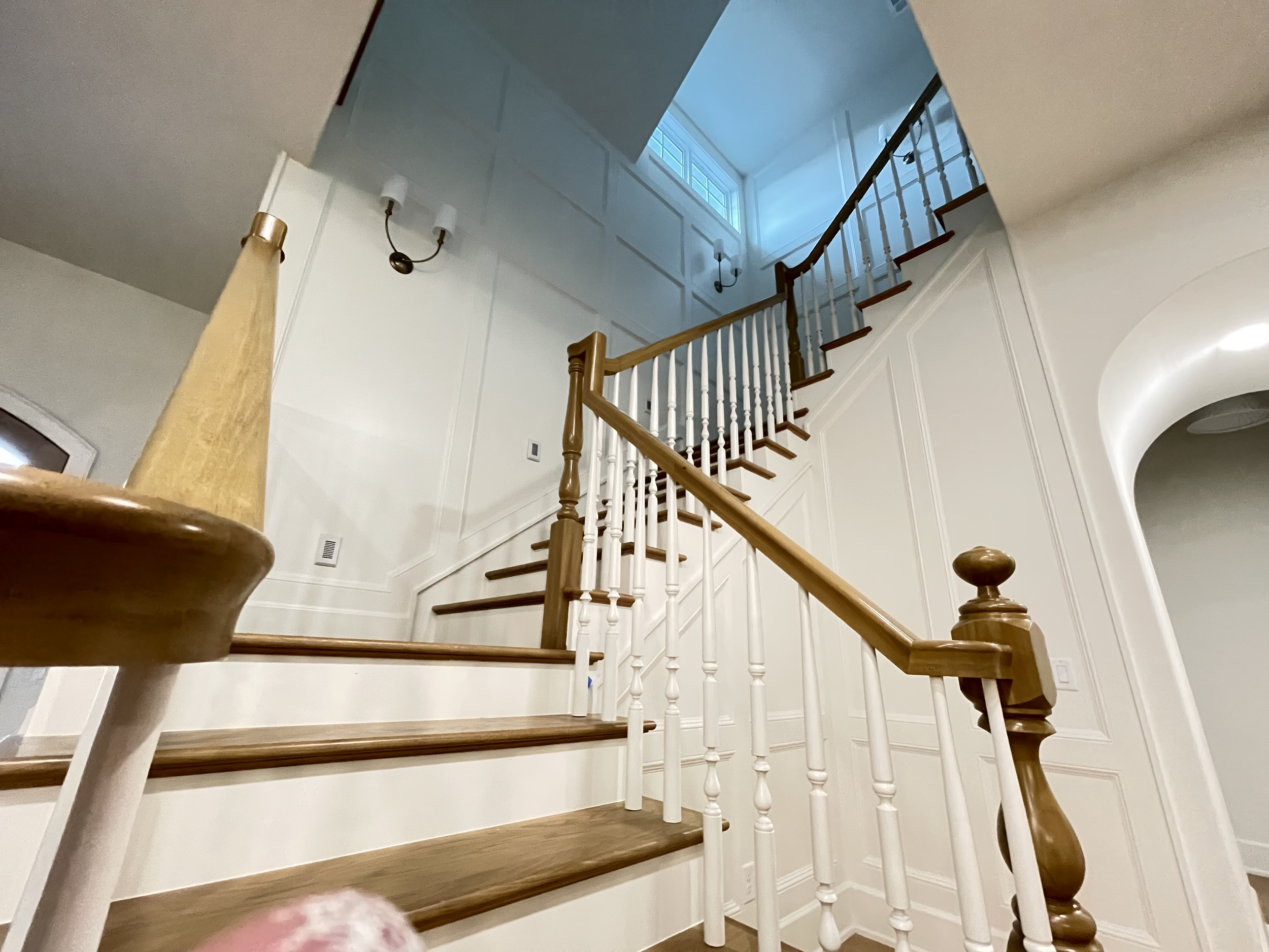 Wooden staircase with white balusters and wooden handrail, leading to an upper floor with a window letting in natural light, interior wall paneling, and a wall-mounted light fixture.