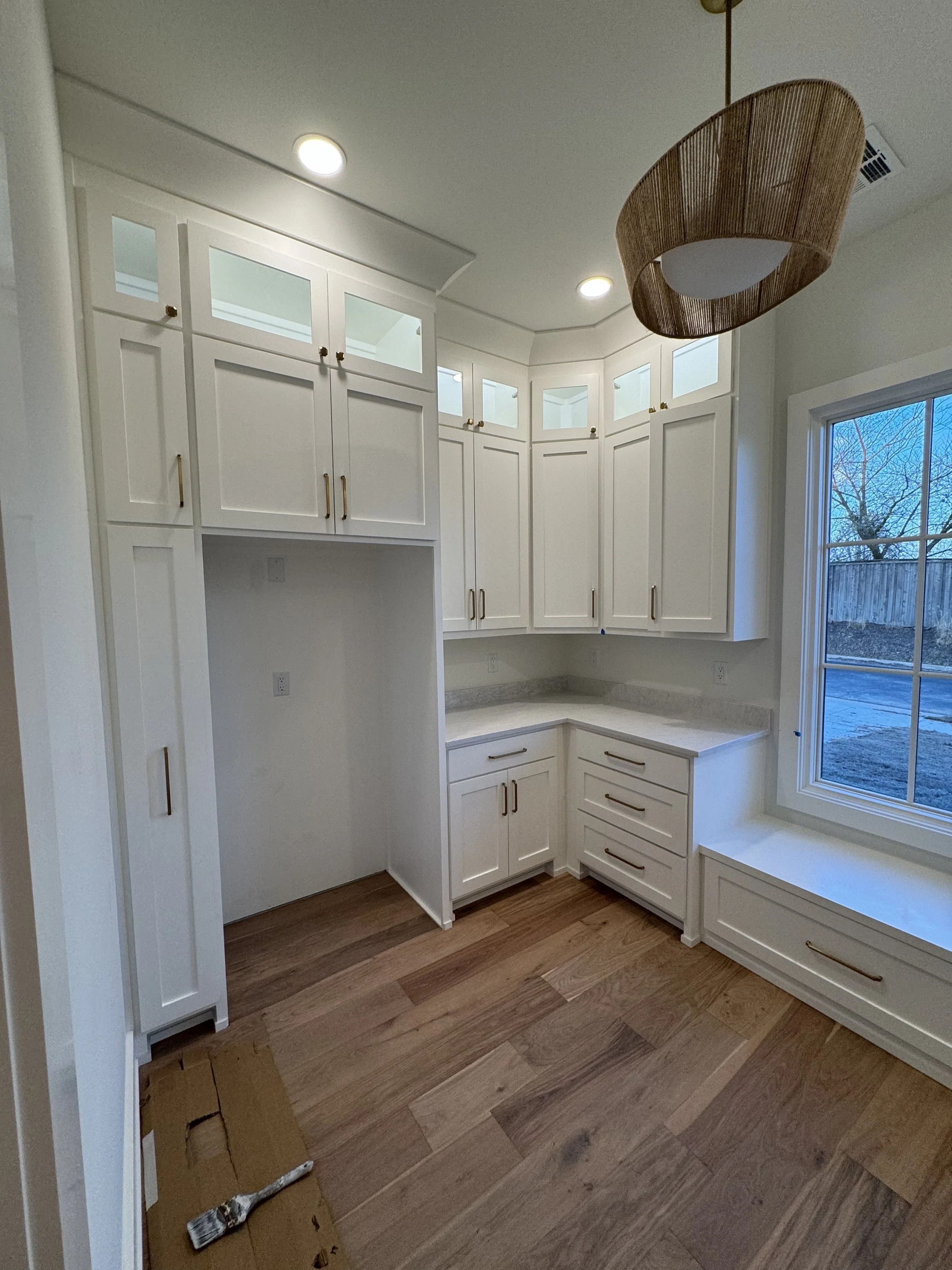 Empty kitchen corner with white cabinets, marble countertops, wood flooring, and a large window showing an outdoor yard.