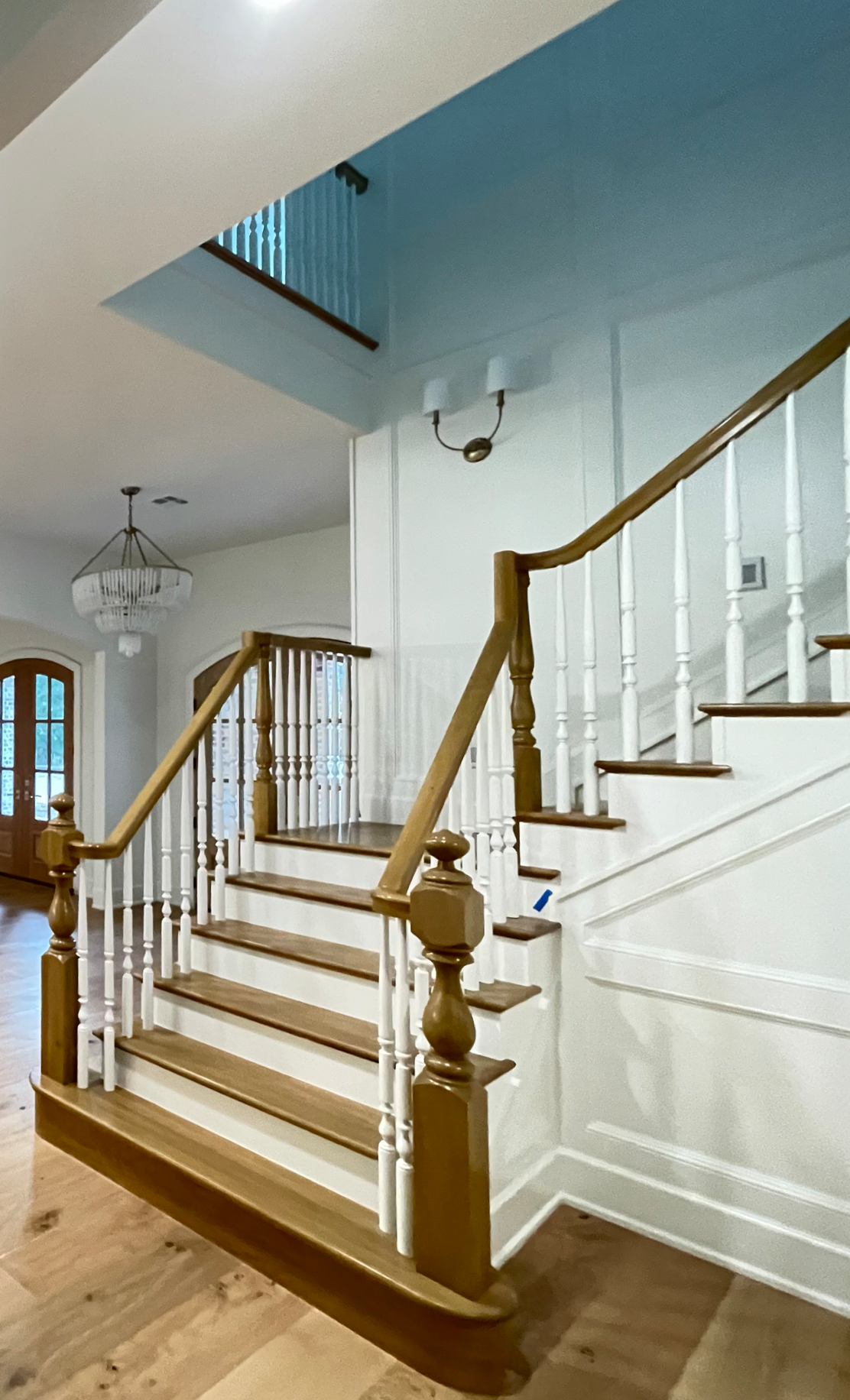 Wooden staircase inside a house with white balusters and light wood handrails, leading to an upper floor with a partial view of a balcony with a wooden railing.