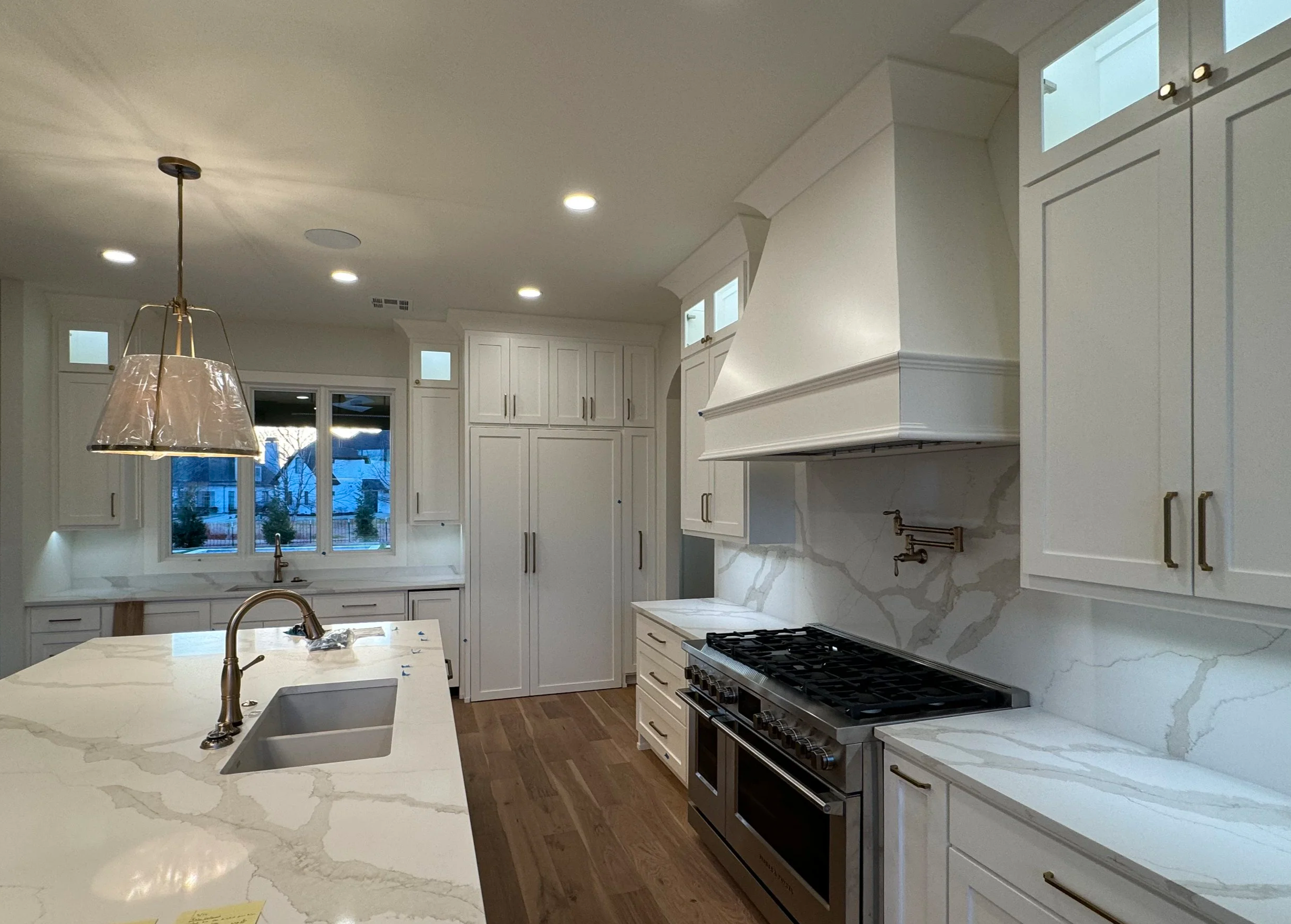 Modern white kitchen with island, gas stove, marble countertops and wood flooring, large window showing outside neighborhood.