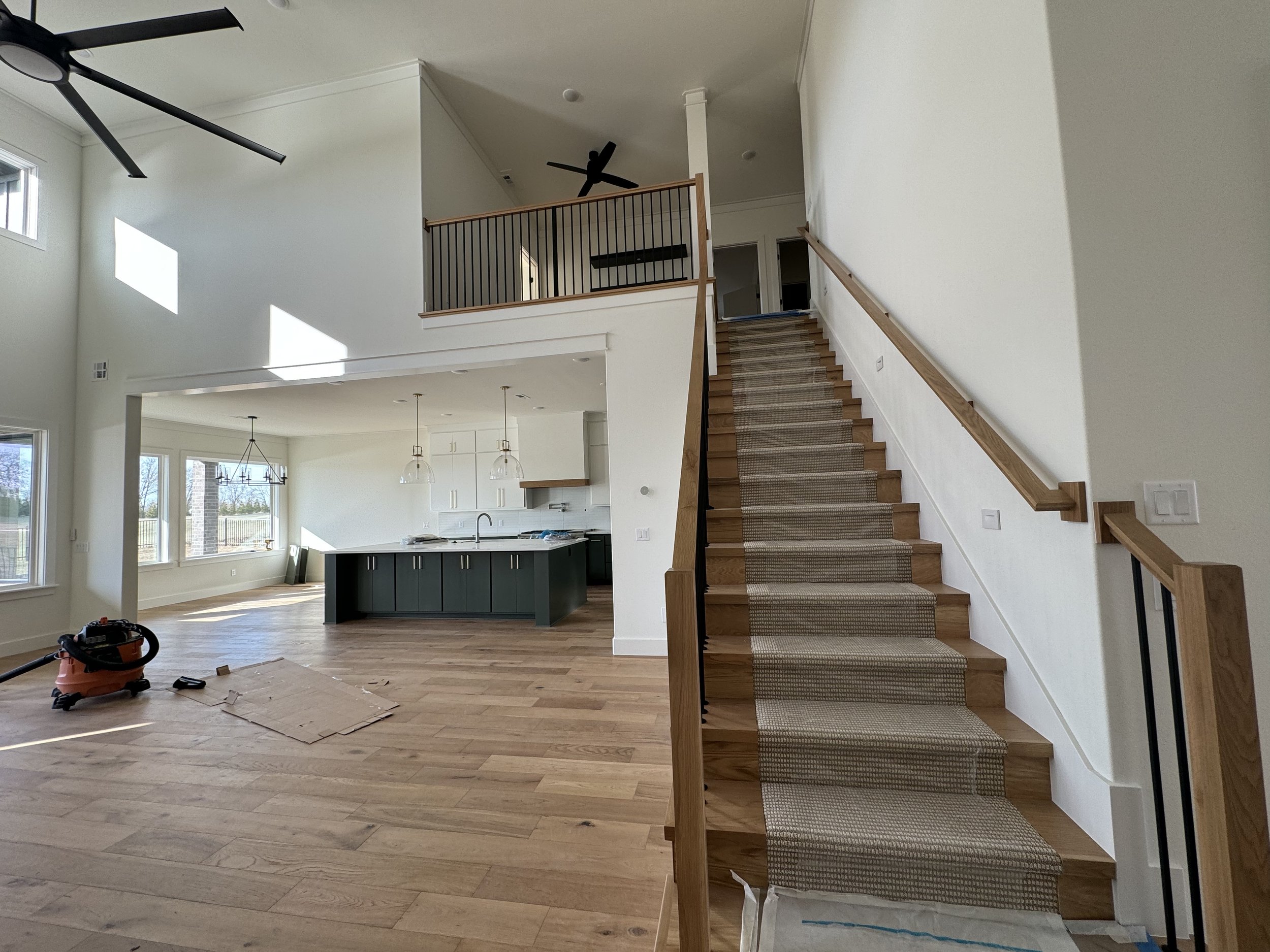 Interior view of a modern house with a staircase, living room, and kitchen area. The staircase has wooden steps and a beige carpet runner. The living room features large windows, light-colored hardwood floors, and a three-light chandelier. The kitche
