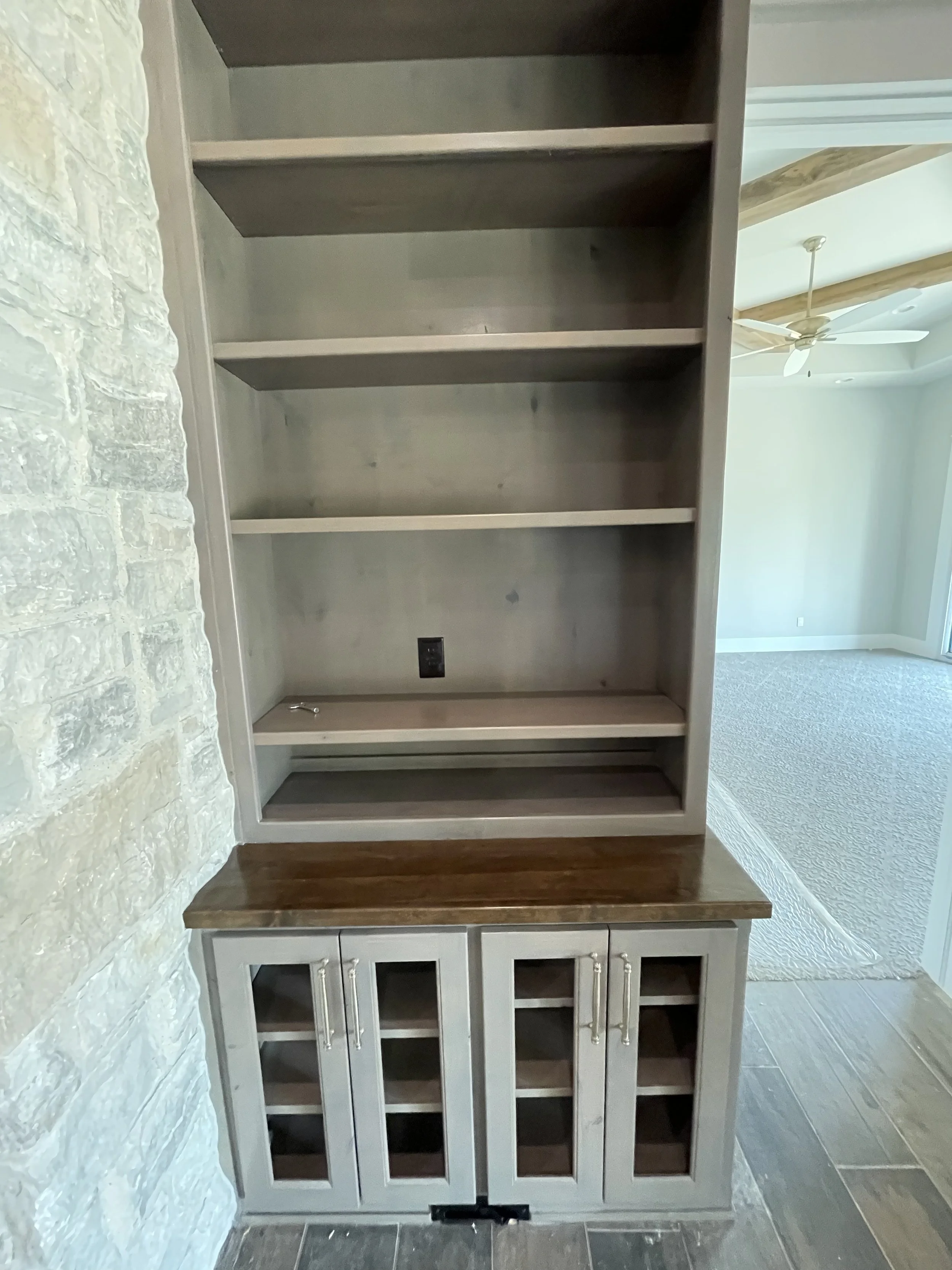 Empty built-in bookshelf and cabinet unit in a room with a stone wall and a view of a ceiling fan and window in the next room.