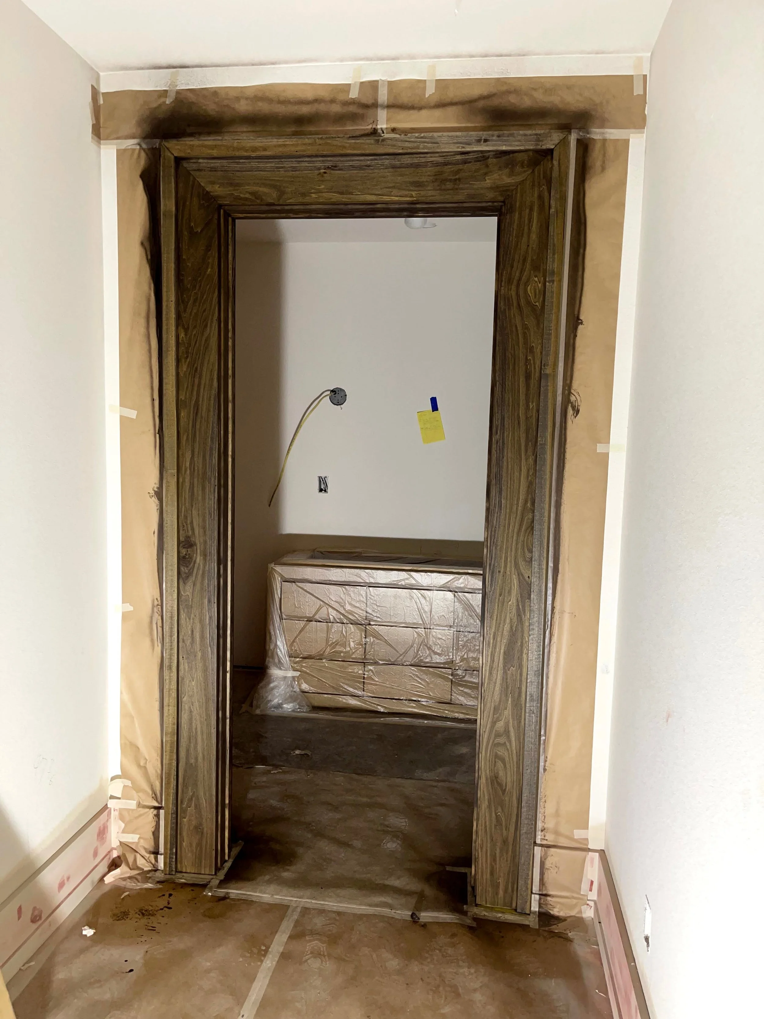 Interior of a room under construction or renovation with a wooden doorway frame, covered with painter's tape, and protective wrapping on nearby furniture. The floor appears unfinished.