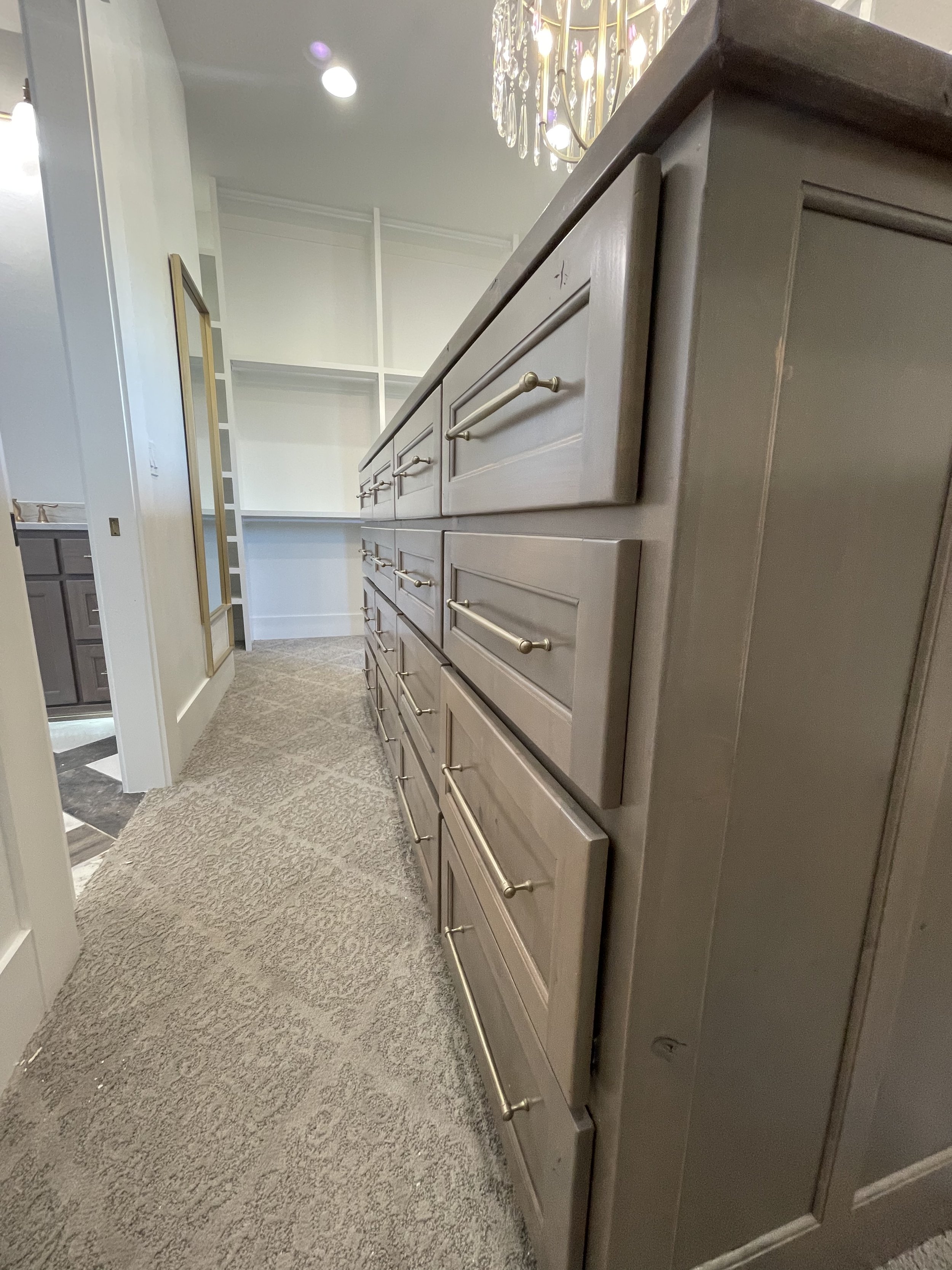 A beige kitchen cabinet with multiple drawers and gold handles, viewed from a low, angled perspective, with a chandelier hanging from the ceiling and open shelving in the background.