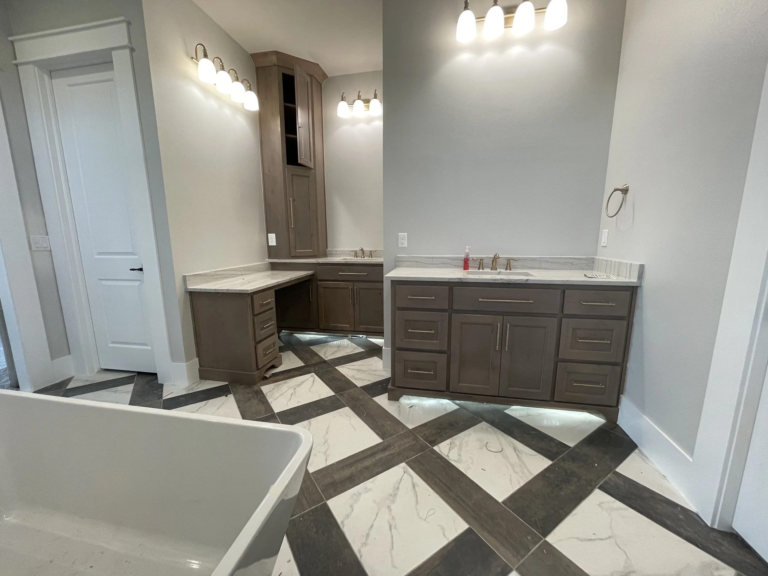 Bathroom with gray cabinets, marble countertops, and black and white geometric tiled floor, with lighting fixtures above the mirrors.
