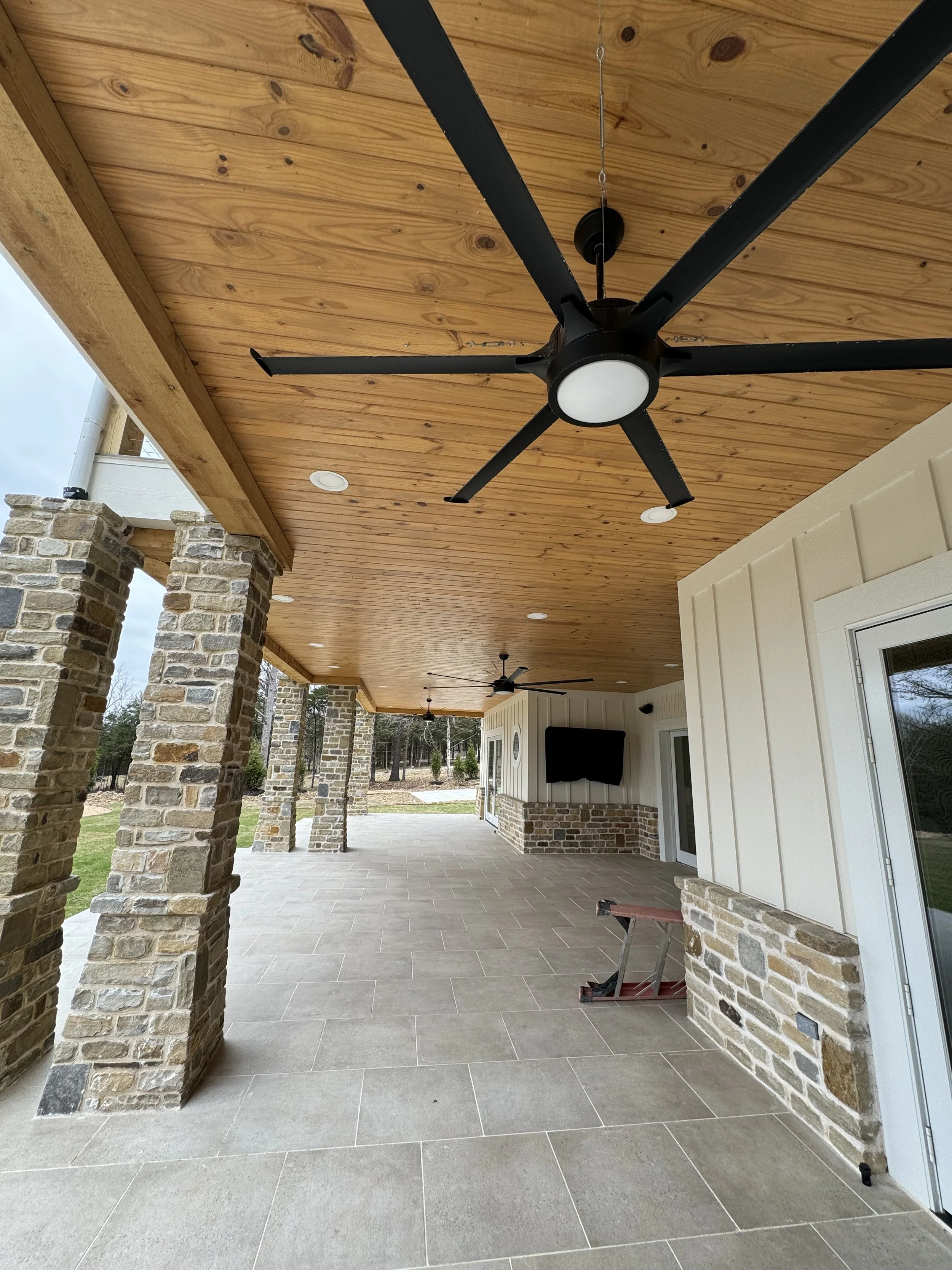 Covered outdoor patio with a wooden ceiling, brick pillars, and ceiling fans. A wall-mounted TV is visible, and a small red table is on the tiled floor. The area is open to a yard with trees.