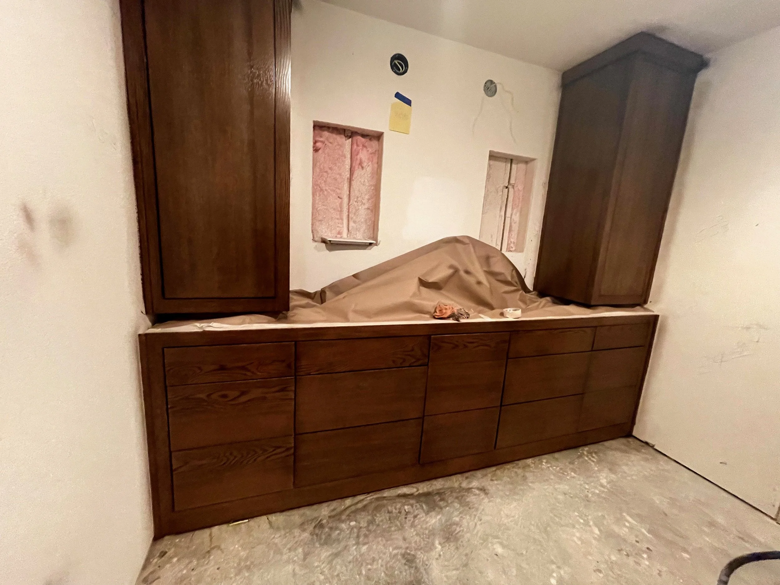 Wooden cabinet with two large top sections and several drawers below, against a white wall with two small, covered windows.
