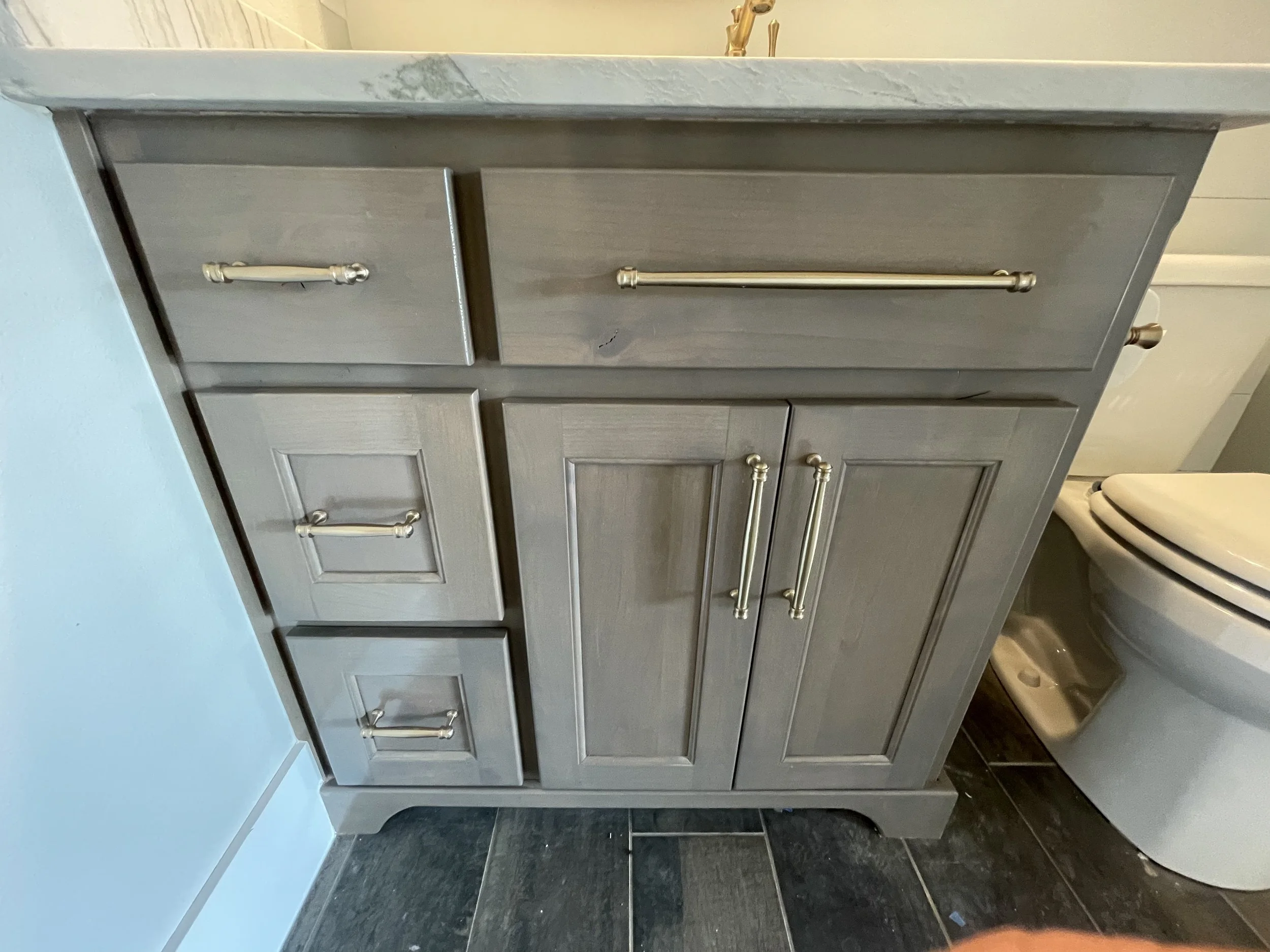 Gray bathroom vanity with four cabinet doors and drawers, gold handles, adjacent to a toilet, with a marble countertop and a gold faucet.