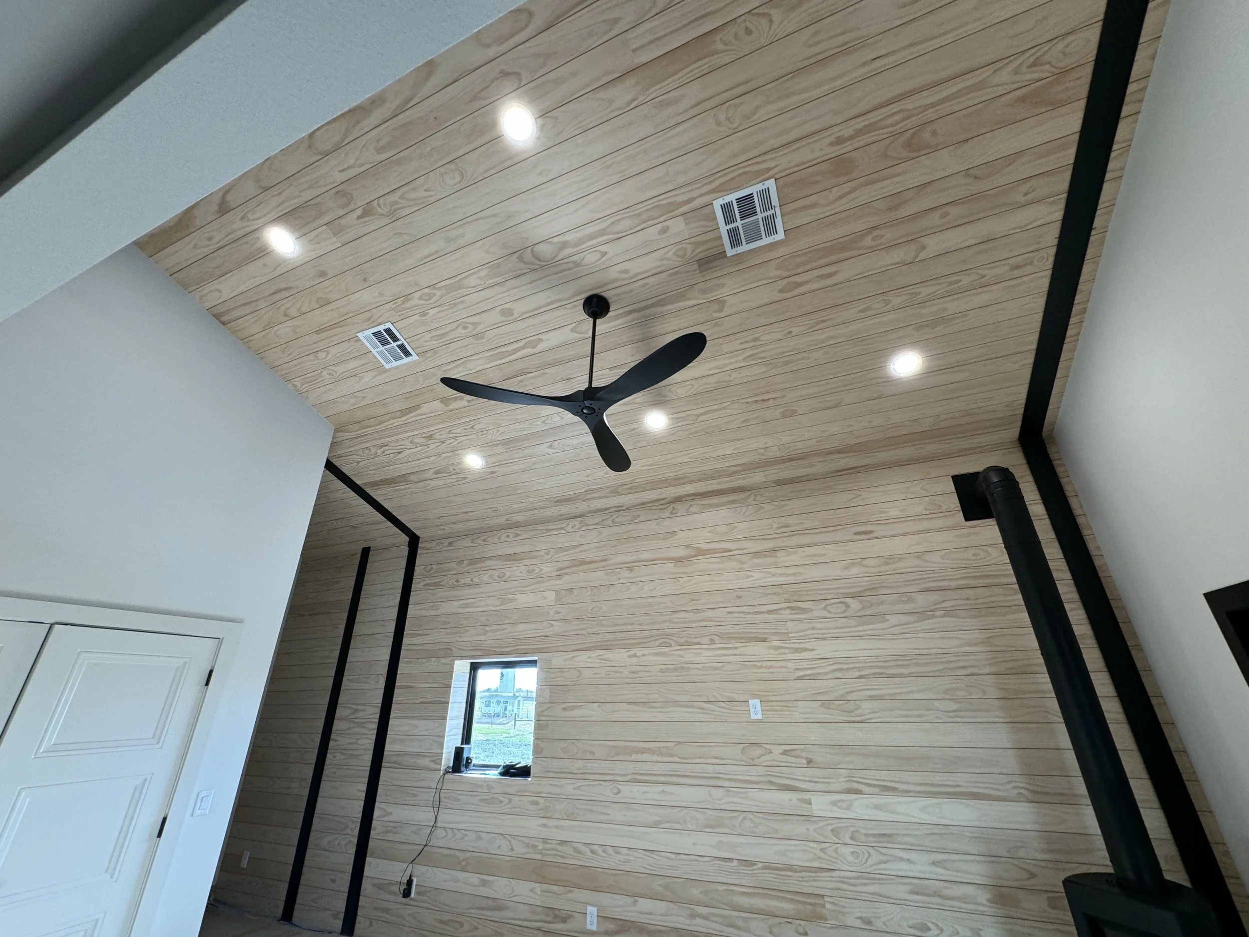 Interior view of a room with a wooden ceiling, black ceiling fan, recessed lights, small window, black metal frame, and black stove pipe.
