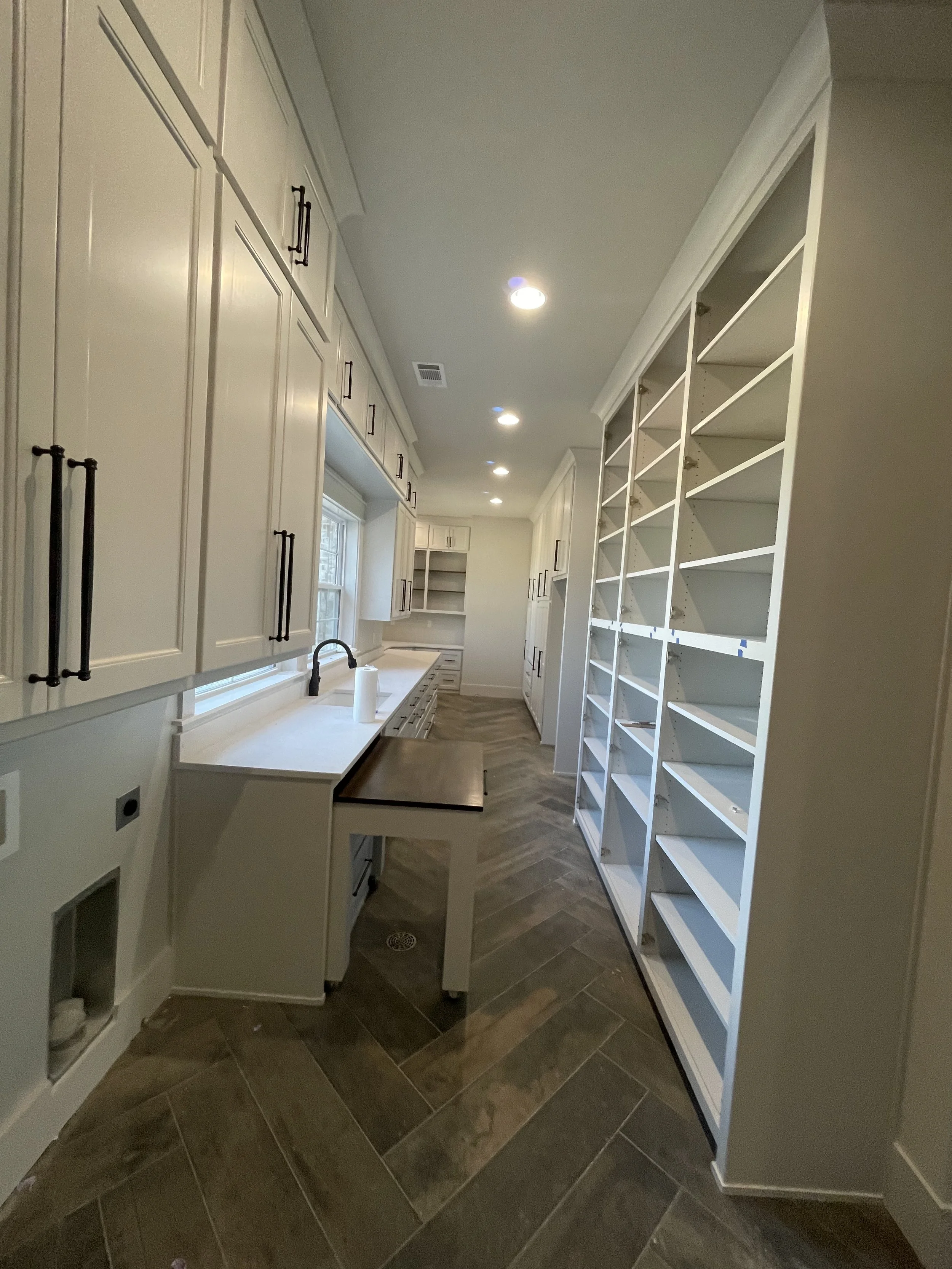 View of a kitchen with white cabinets, a white countertop, a window above the sink, and large empty white shelving units.