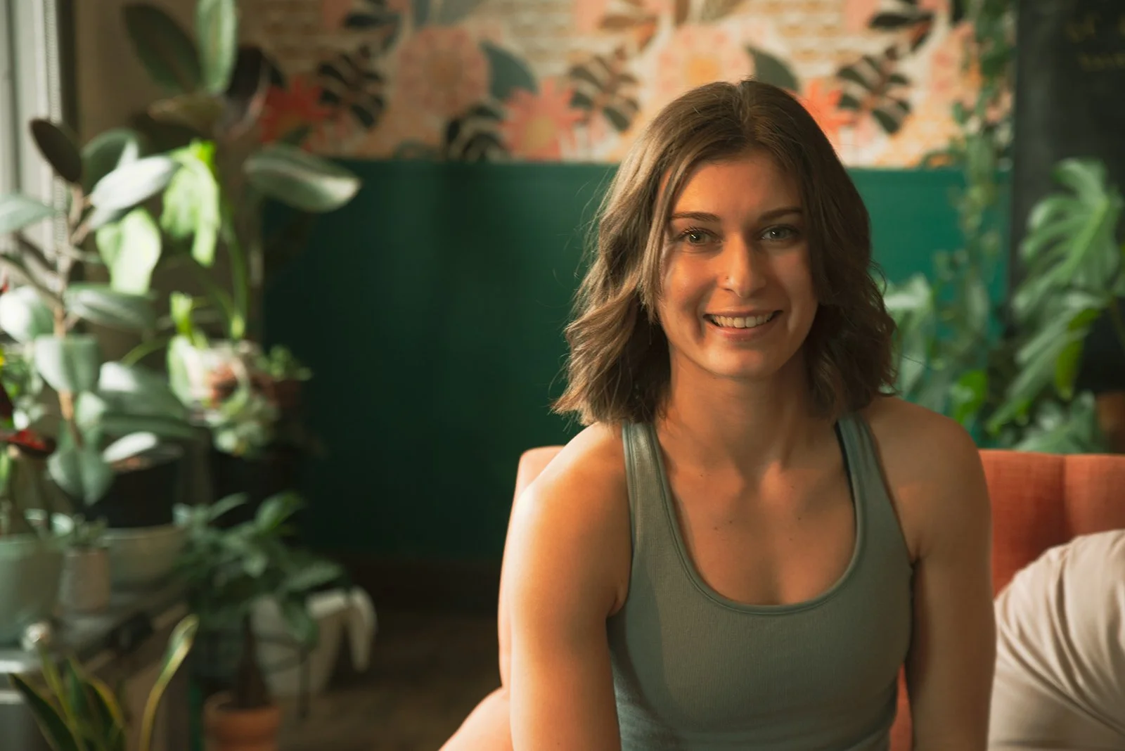 A young woman with shoulder-length wavy brown hair, smiling, sitting indoors in a room filled with green plants, wearing a gray tank top.