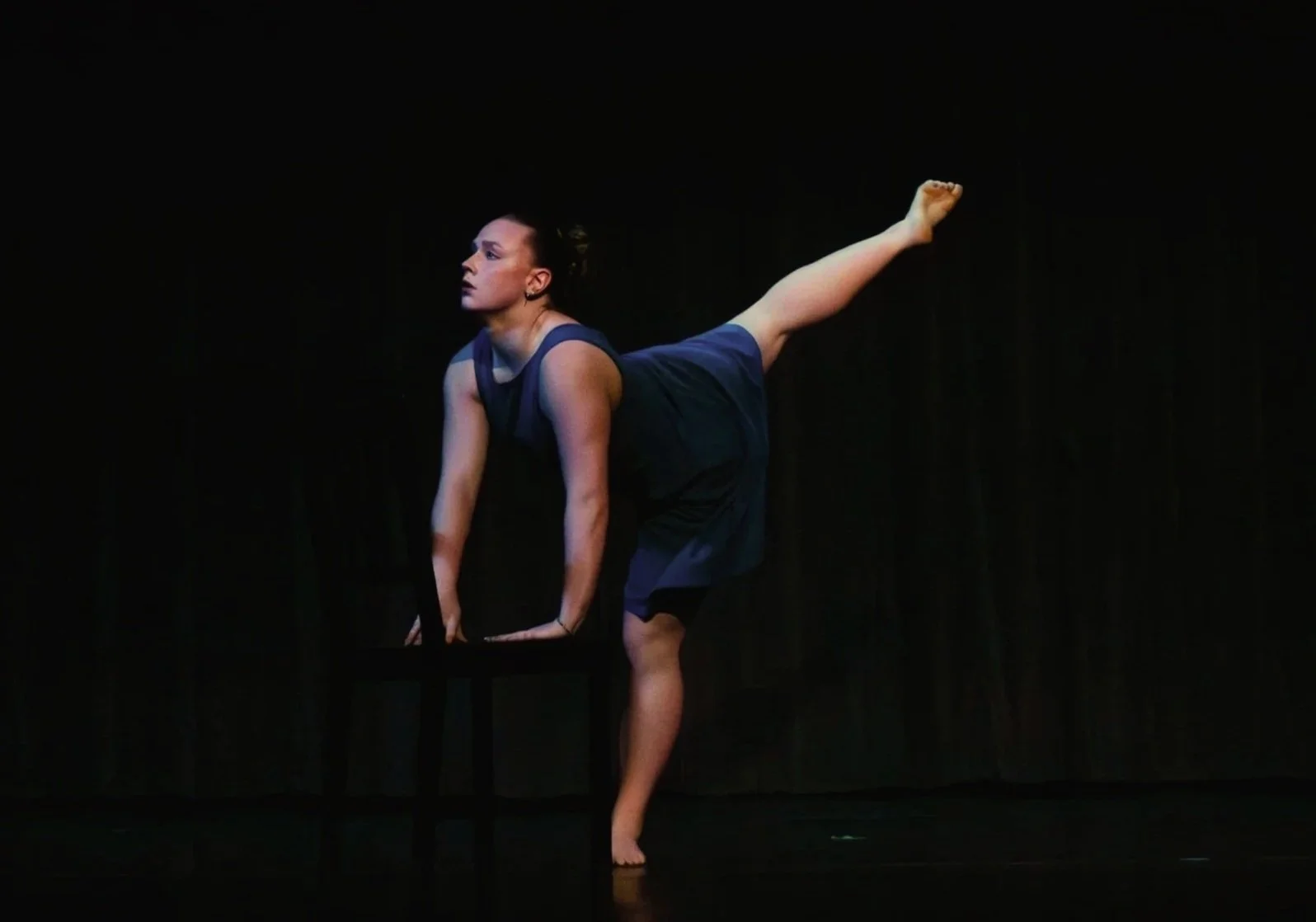 A woman performing a dance move on stage, balancing on her hands with one leg extended back, dressed in a dark blue dress against a dark background.