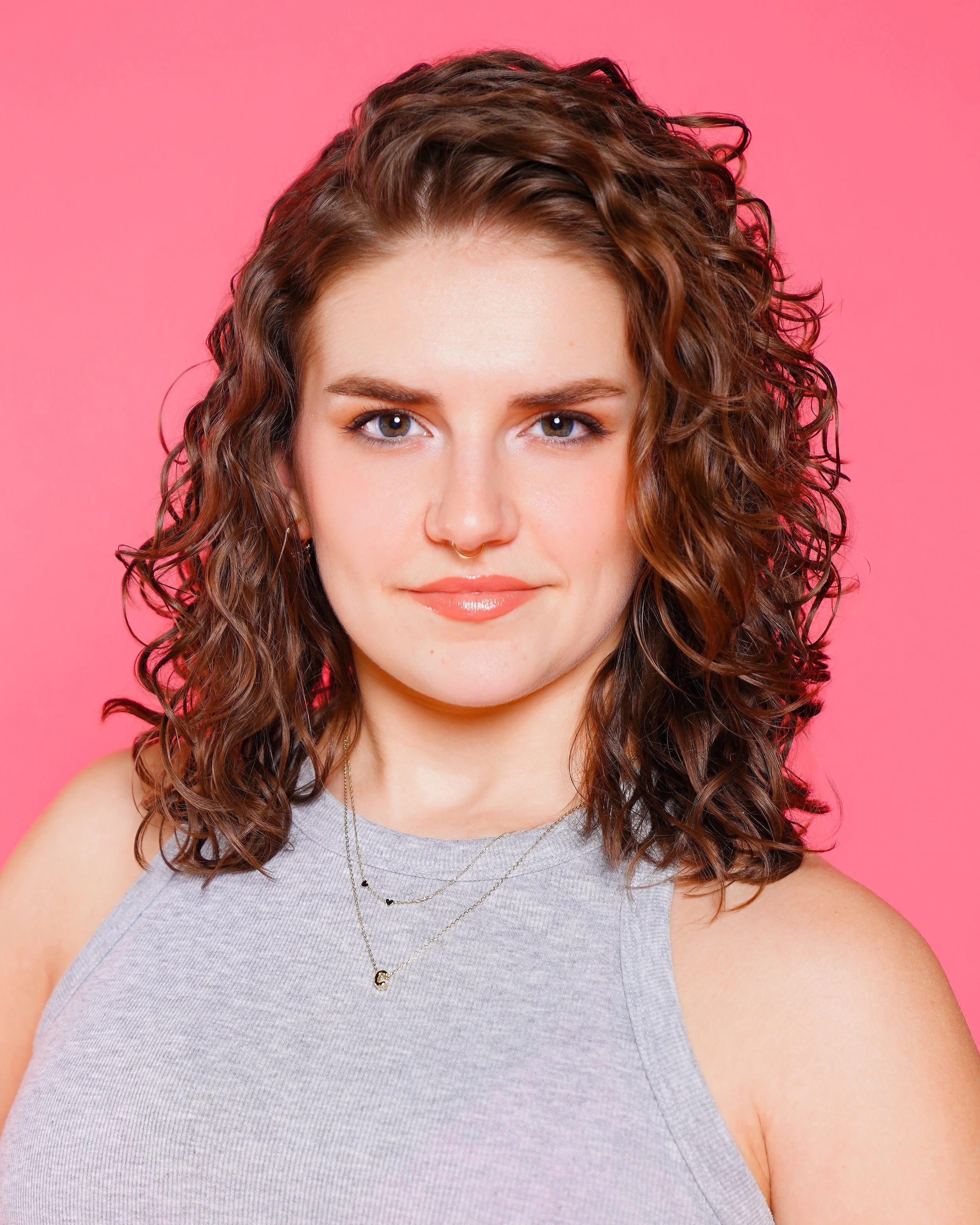 Close-up of a woman with curly brown hair and blue eyes, wearing a gray tank top and layered necklaces, against a pink background.