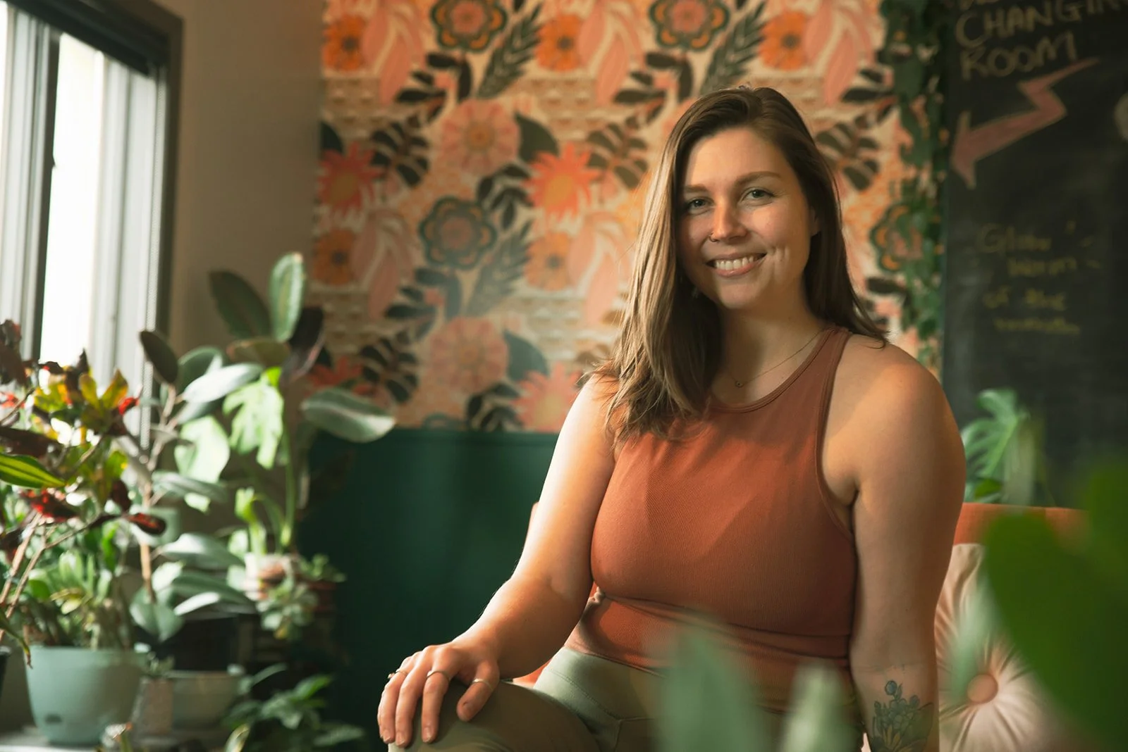 A woman with brown hair smiling while sitting in a room with plants and a floral wallpaper background.