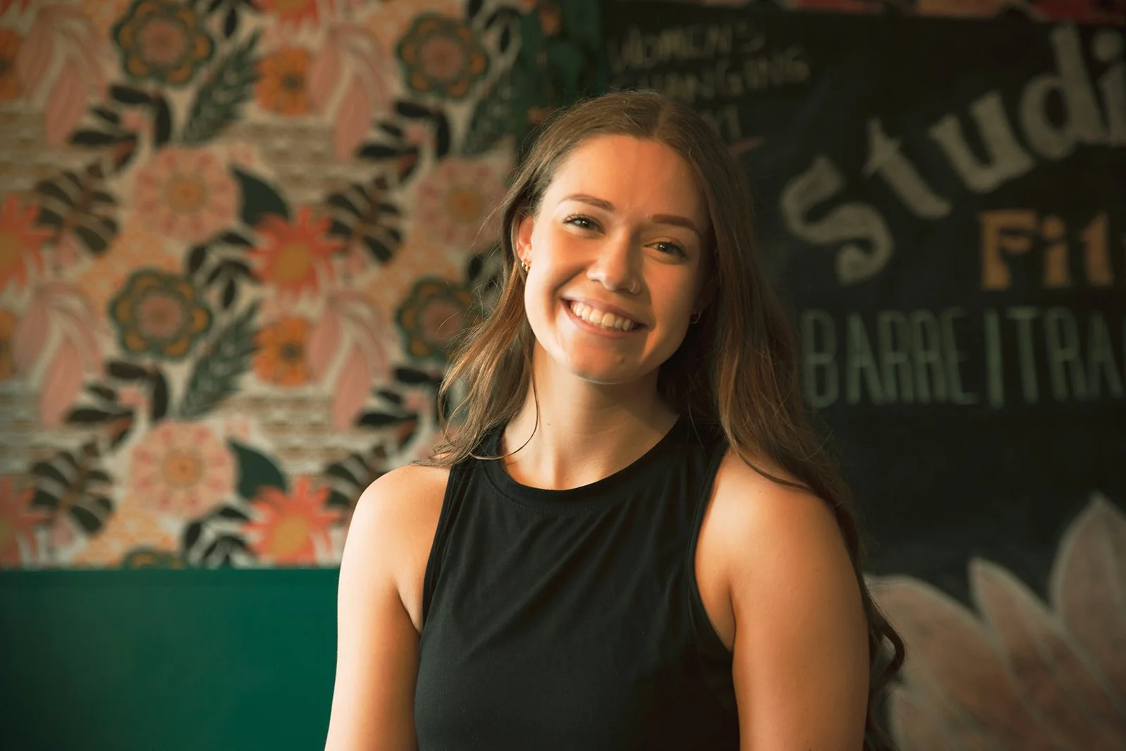 A young woman with long brown hair smiling, wearing a sleeveless black top, in front of a floral-patterned wall and a chalkboard menu.