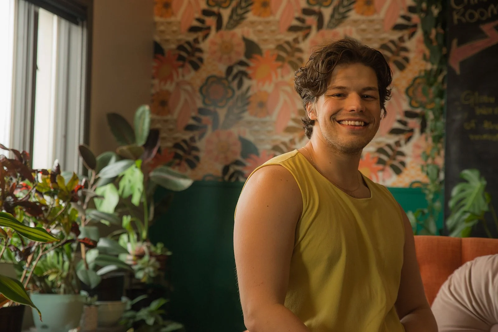 A young man with curly brown hair wearing a yellow sleeveless shirt sitting indoors, smiling at the camera, with a floral wallpaper and plants in the background.