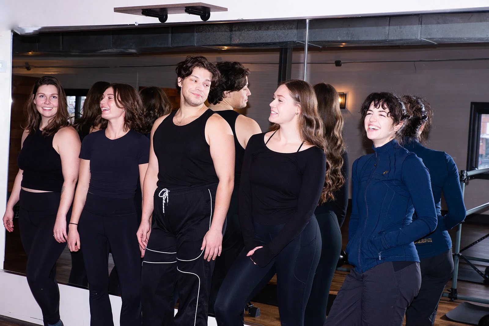 A group of young people in athletic clothes are gathered in a fitness studio, some are stretching and chatting.