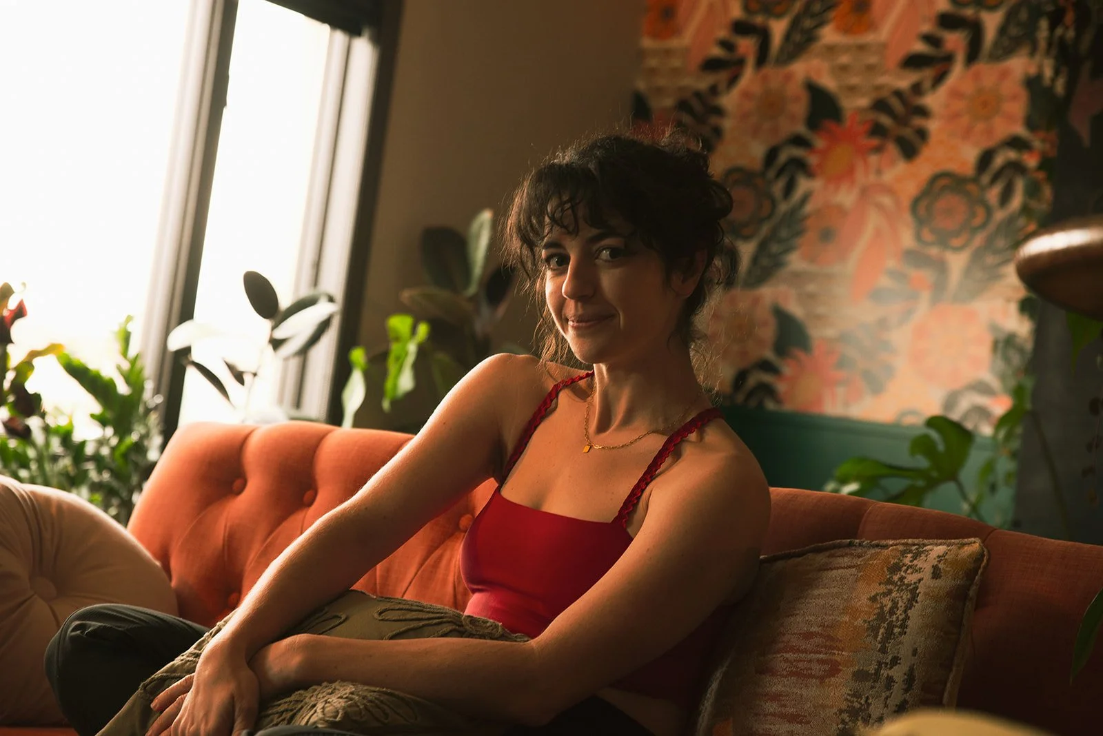 Woman in red workout attire smirking at camera while seated in front of plants and floral wallpaper