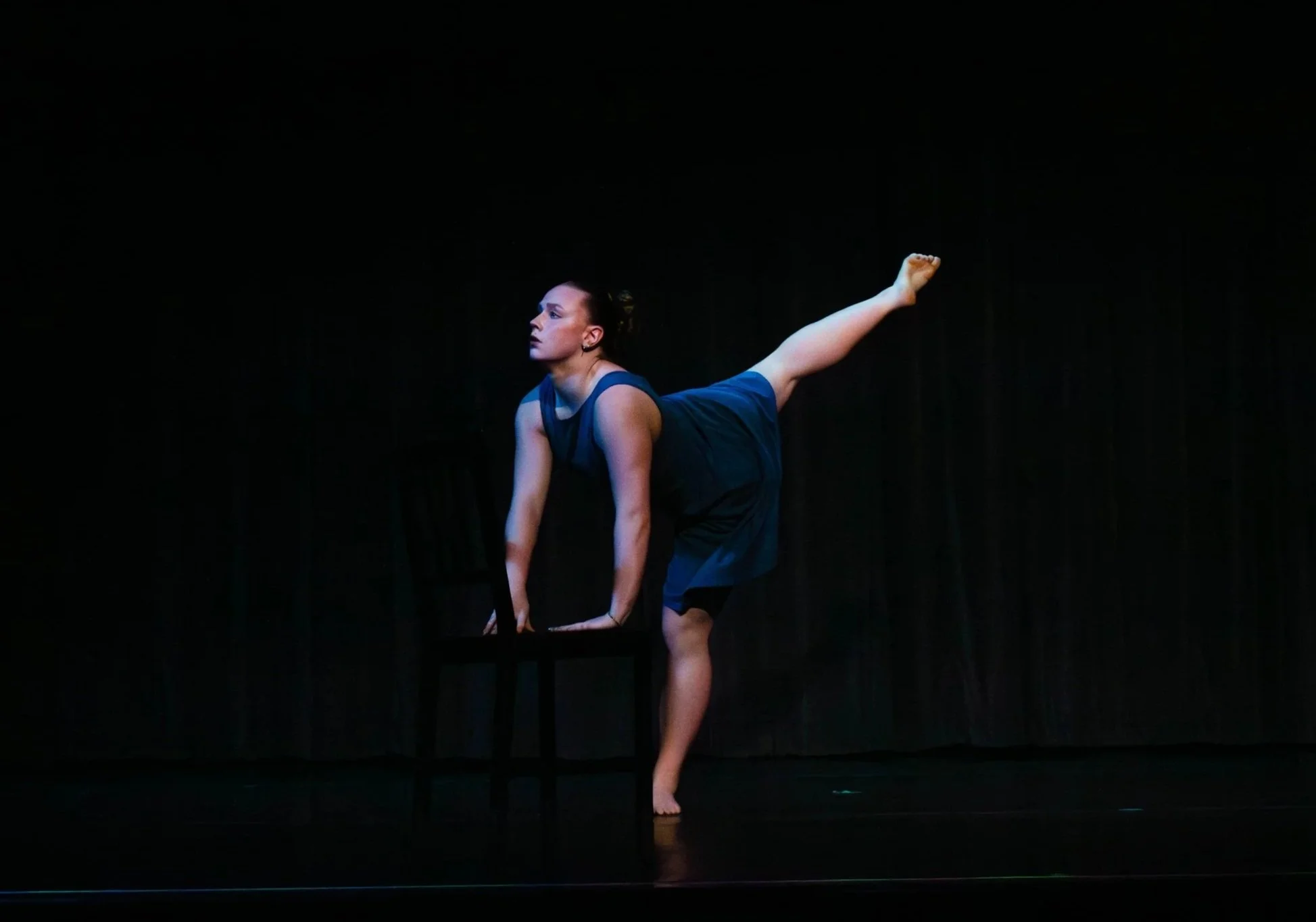 A dancer in a blue dress practicing a pose on stage, with a dark background.
