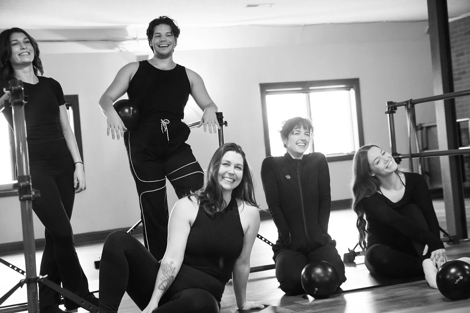Five women in workout clothes smiling and posing in a fitness studio with exercise equipment, including kettlebells and a ballet barre, in the background.