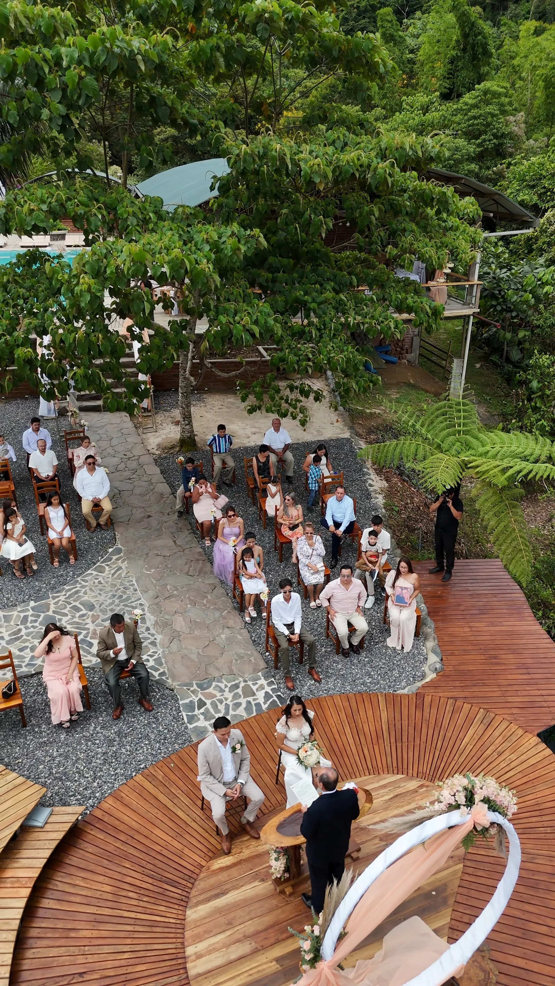 Una ceremonia de boda al aire libre con invitados sentados en sillas de madera en un entorno natural con árboles y vegetación. La pareja de novios está en un altar circular de madera decorado con flores, frente a un oficiante.