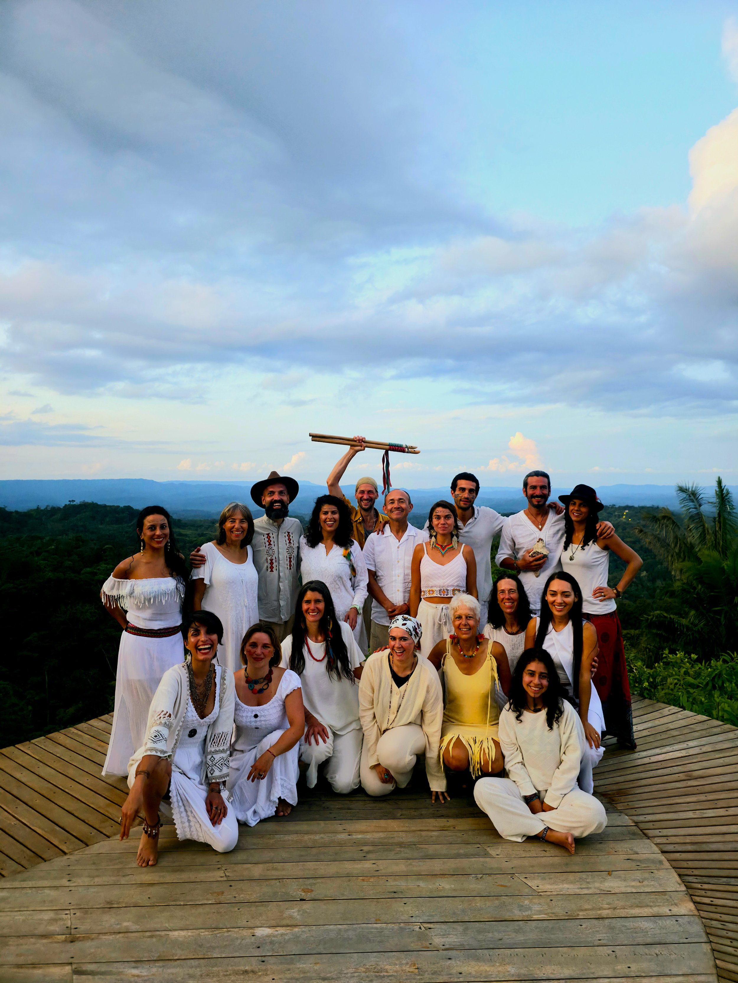 Grupo de personas vestidas con ropa blanca en un escenario al aire libre, con un paisaje natural y cielo nublado en el fondo, posando y sonriendo.