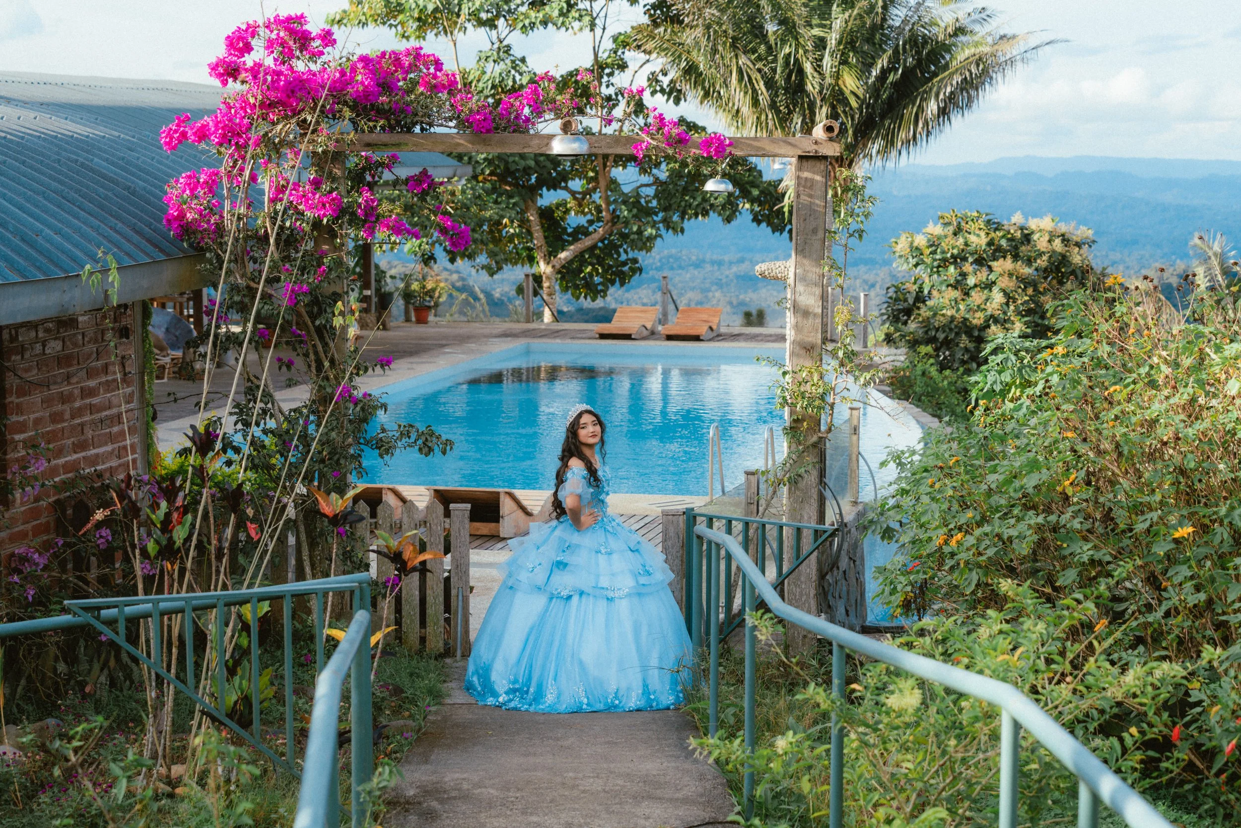 Jovencita quinceñera vestida con un vestido azul princesa en un camino que conduce a una piscina, rodeada de plantas y flores, con árboles y montañas de fondo.