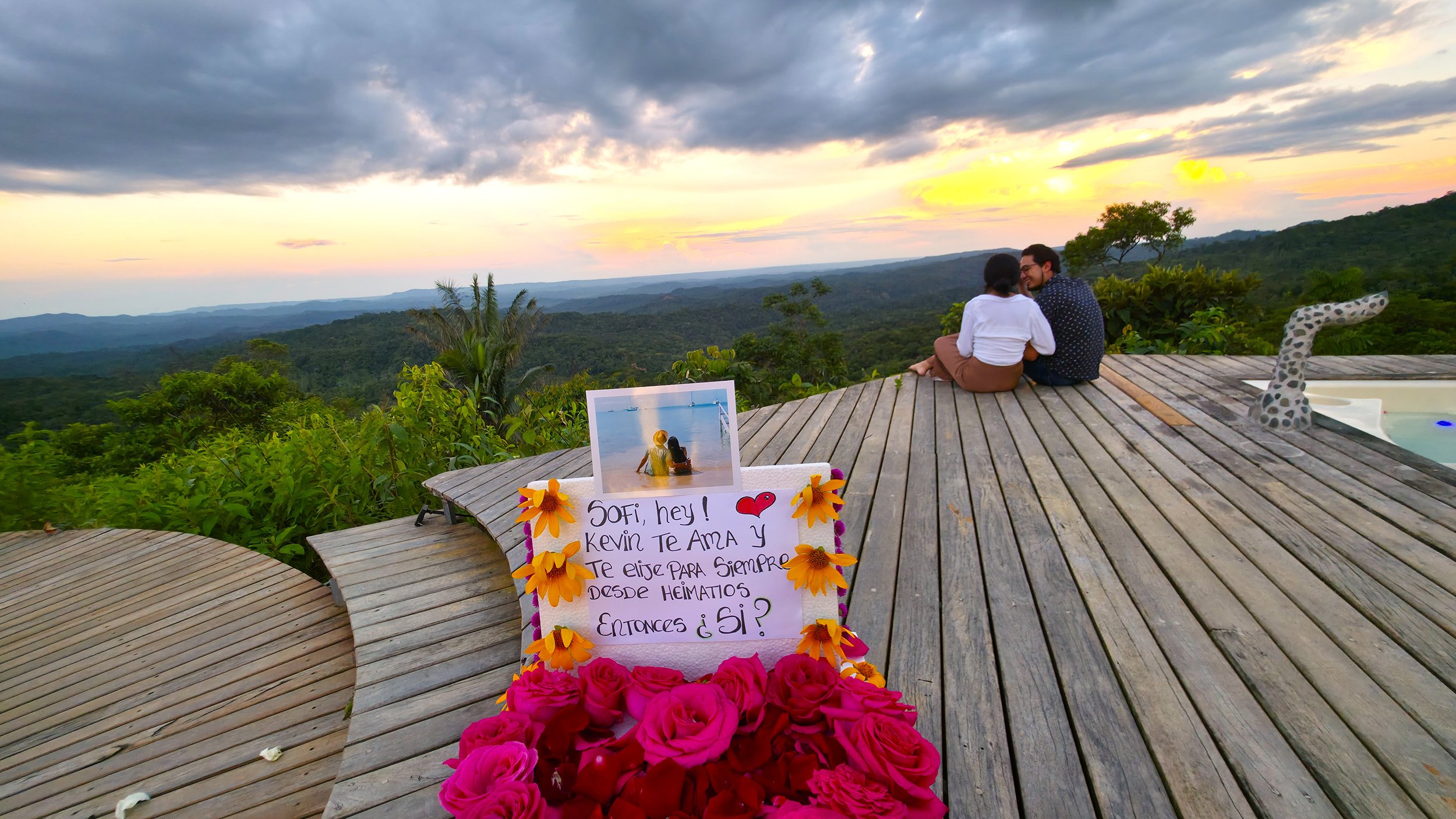Pareja sentada en una terraza de madera con vista a un paisaje de montañas y árboles durante el atardecer, con una silla en forma de jirafa en la esquina derecha y una cartulina con mensaje de amor decorada con flores en primer plano.