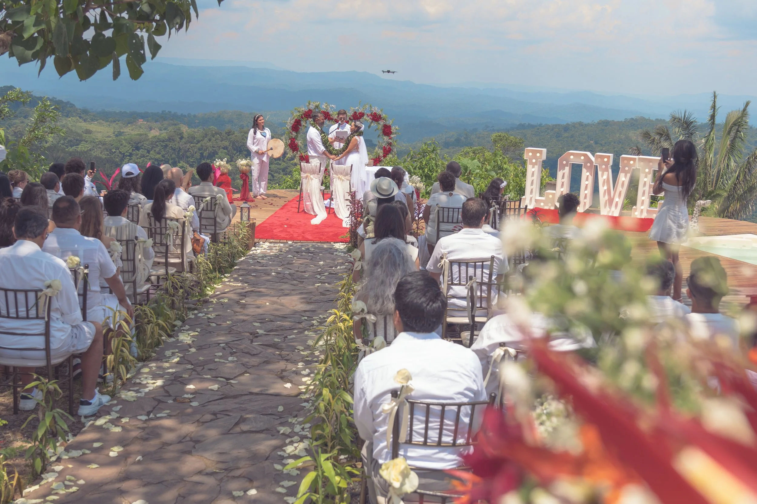 Ceremonia de boda al aire libre en un escenario en las montañas, con invitado y pareja en el altar, decoraciones florales y fondo de naturaleza y cielo.