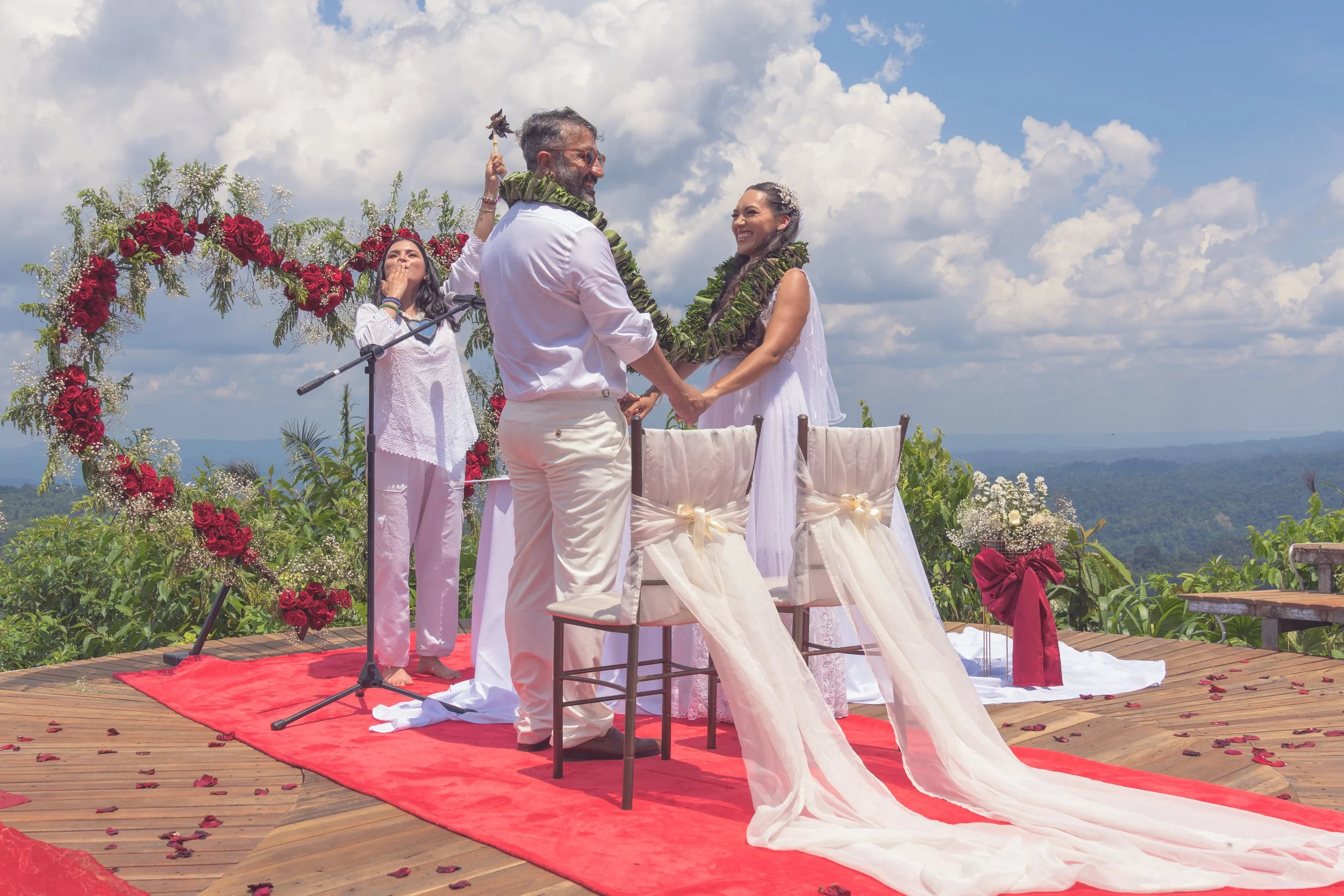 Pareja casándose en una ceremonia al aire libre con fondo de montañas y cielo con nubes, decorada con flores rojas y blancas, con un oficiante y una maestra de ceremonia, en un escenario de madera con alfombra roja.
