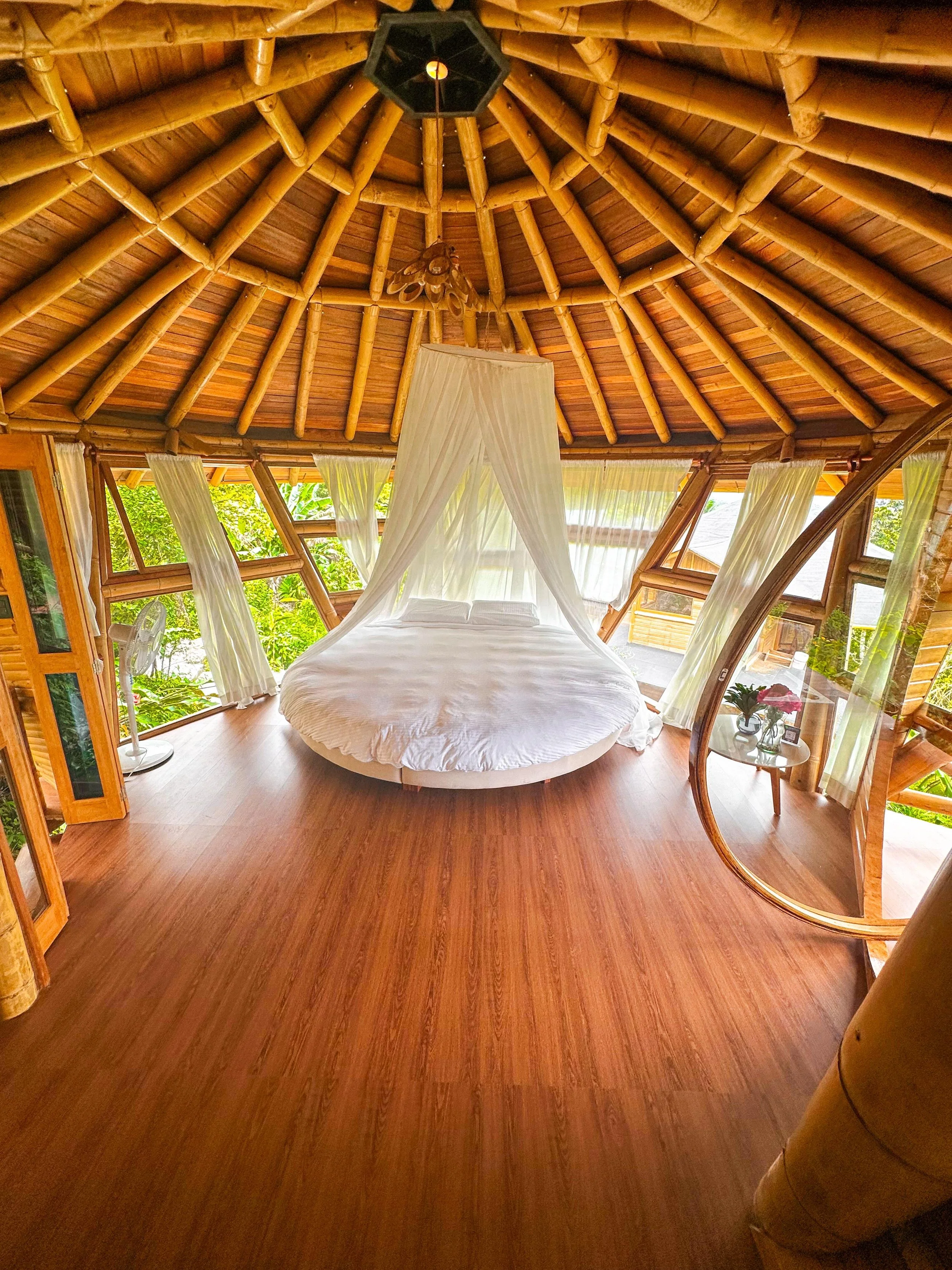 Habitación rústica con techo de bambú, cama redonda con canopy, cortinas blancas, ventanas de madera abiertas y vista a la vegetación exterior.