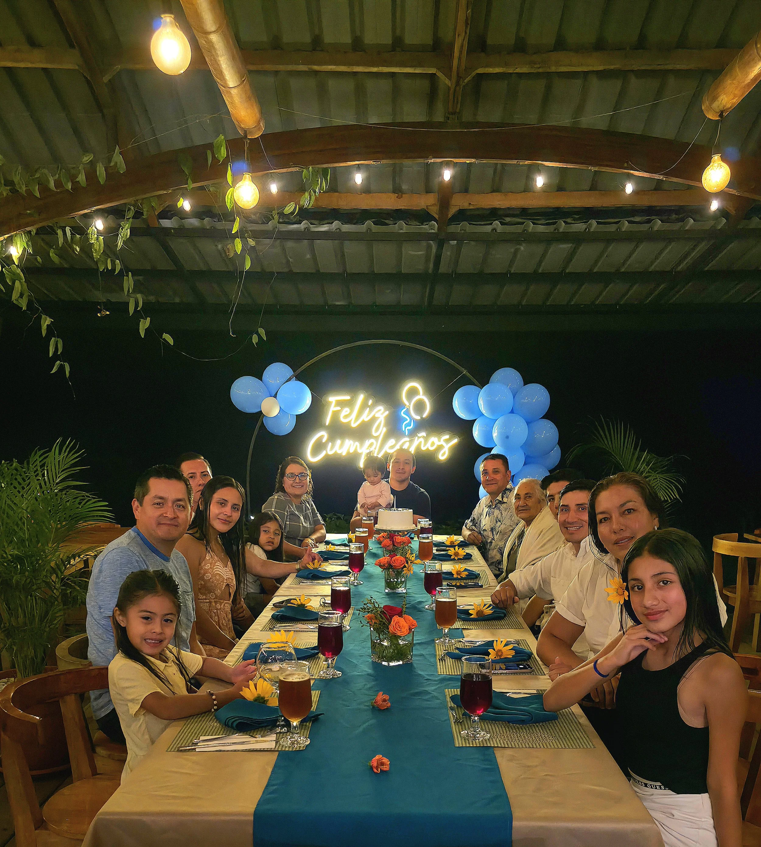 Grupo de personas celebrando un cumpleaños en una mesa larga decorada con flores y bebidas, con un cartel que dice 'Feliz Cumpleaños' y globos azules en el fondo