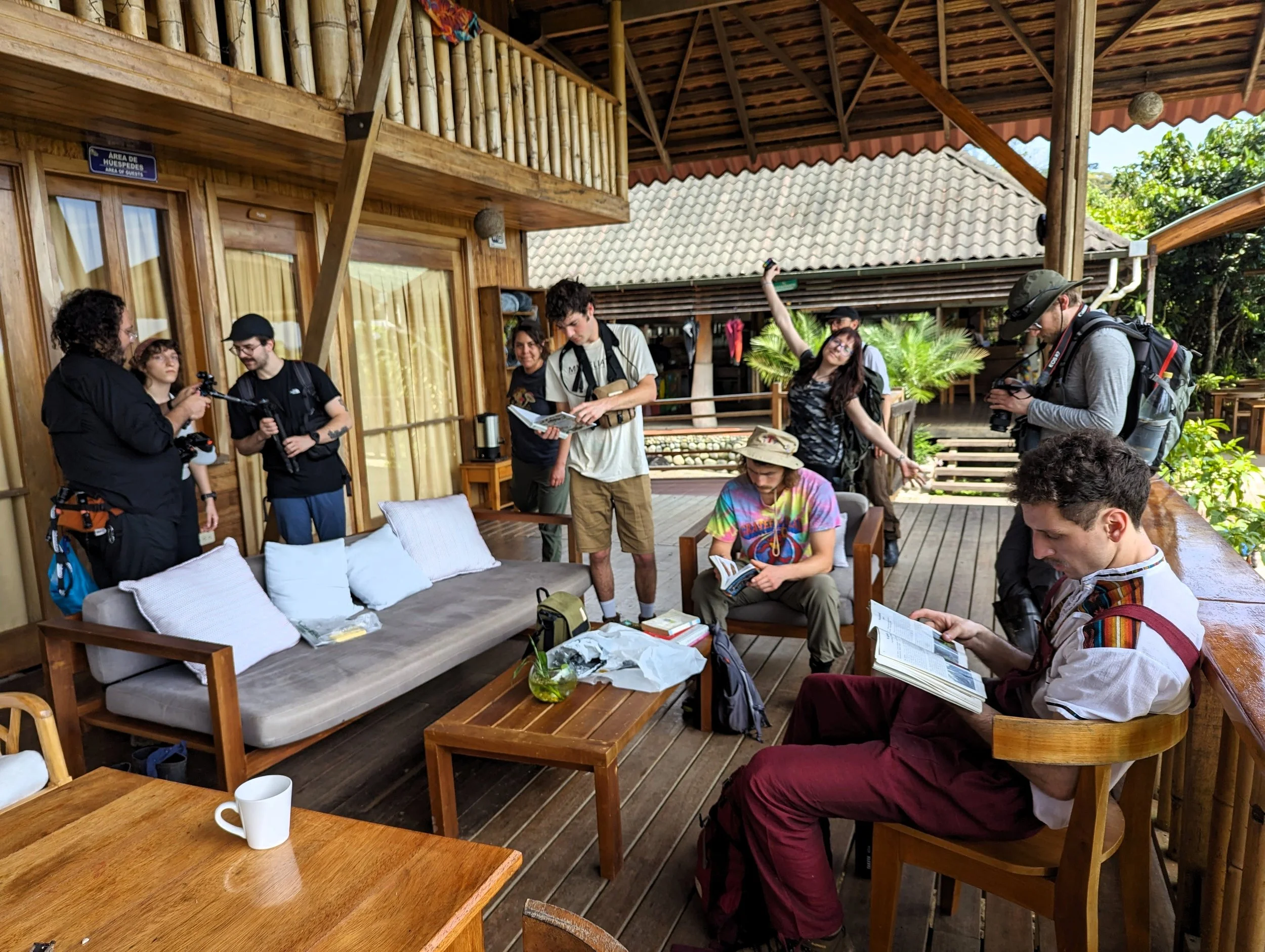 Grupo de personas en una terraza de la casa grande, con algunos leyendo libros y otros tomando fotos o hablando, en un entorno natural con plantas y árboles verdes.