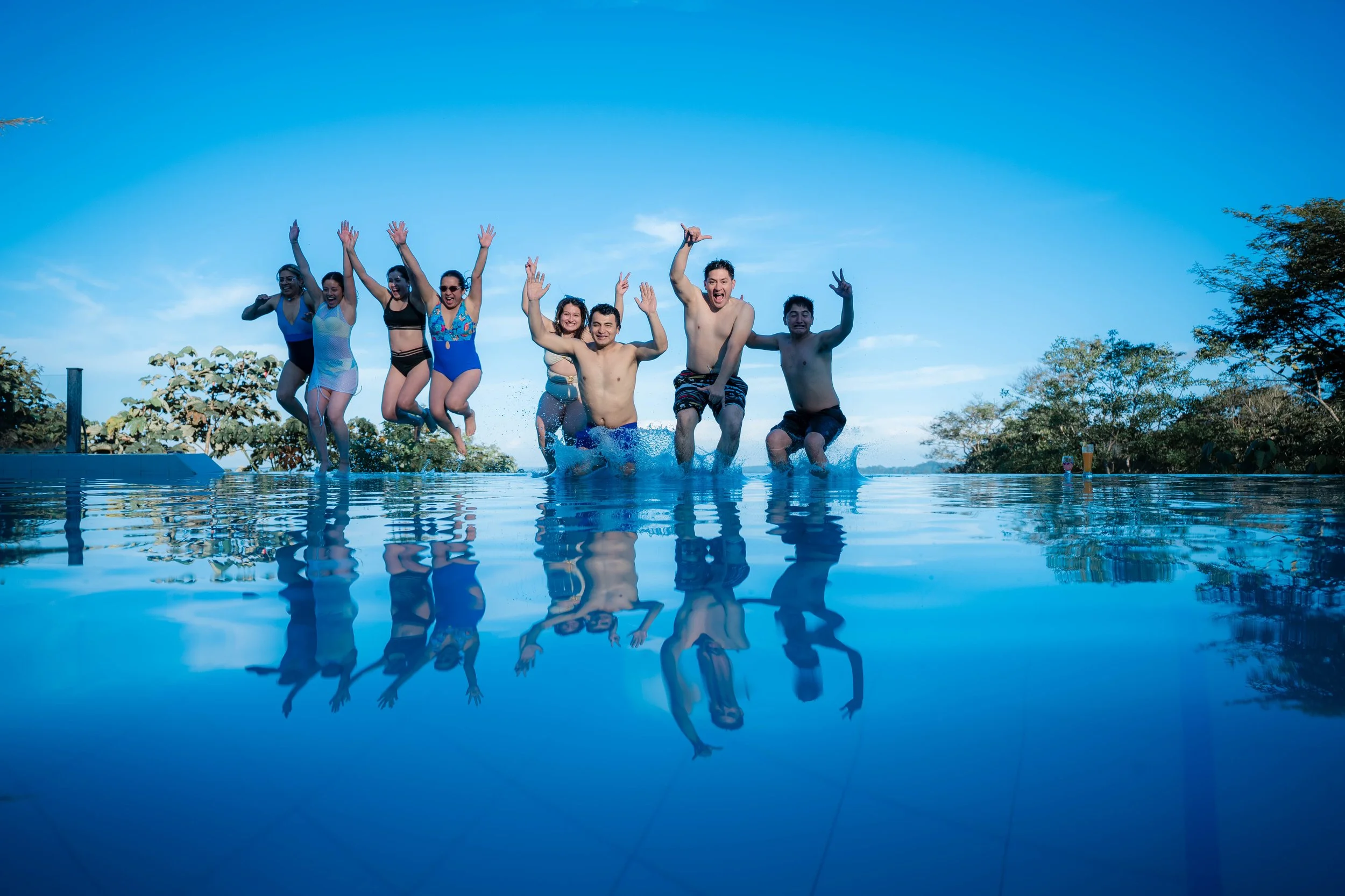 Grupo de personas saltando en una alberca, reflejándose en el agua, con un cielo azul y árboles en el fondo.