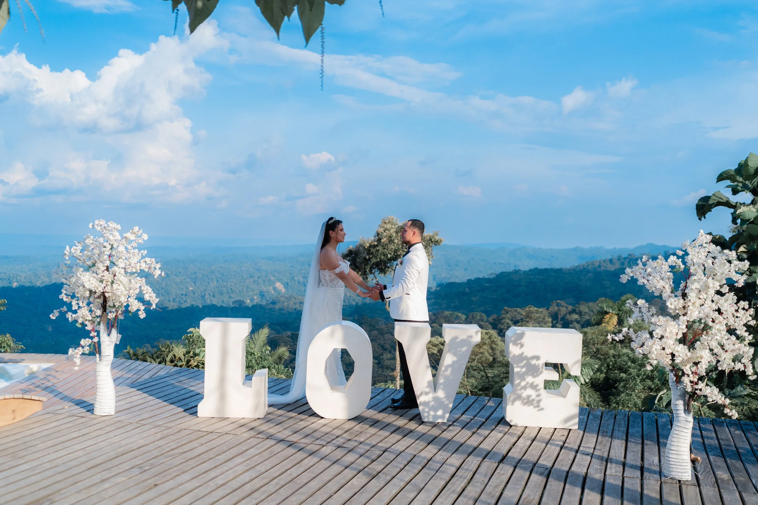 Captura de un momento mágico durante una boda de destino. Los novios lucen elegantes trajes blancos en un mirador de madera con una vista panorámica espectacular hacia la selva.