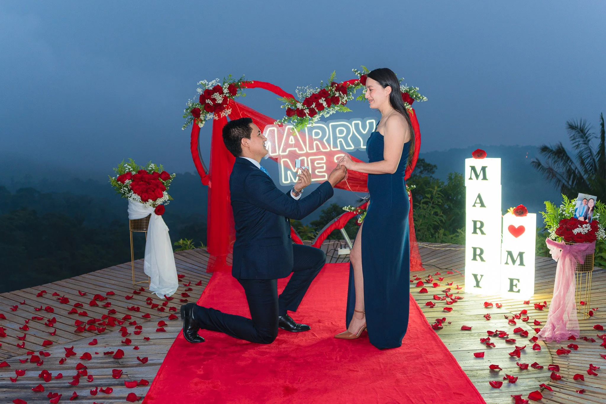 Una pareja en una propuesta de matrimonio en el mirador con flores rojas y un letrero de neón que dice 'Marry Me'. La mujer viste un vestido largo azul y el hombre un traje oscuro, él está de rodillas sosteniendo la mano de ella.
