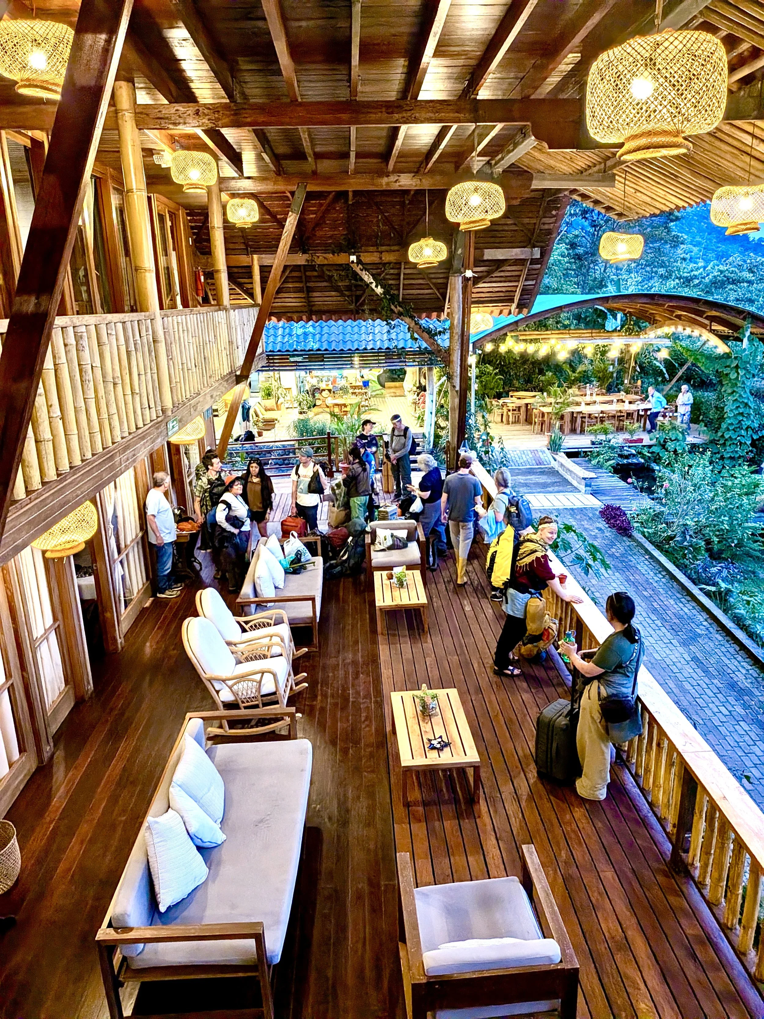 Salón con estructura de madera, plantas y luz calida, con personas esperando y sentadas en área de descanso y pasillo, decorado con lámparas de mimbre y muebles de madera y cojines blancos.