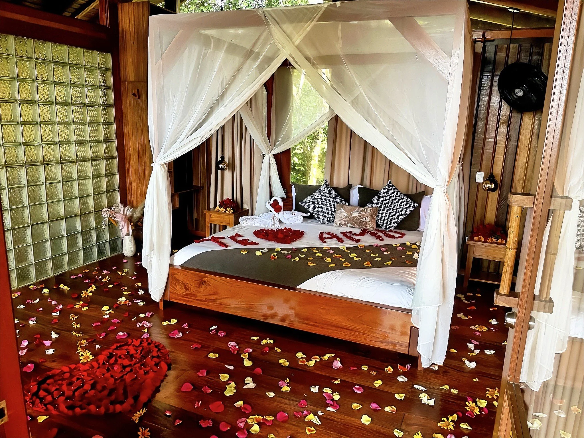 Una cama decorada con pétalos de rosa en forma de corazón y la palabra 'LOVE' en la cama, en una habitación con cortinas blancas, paredes de madera y decoración romántica.