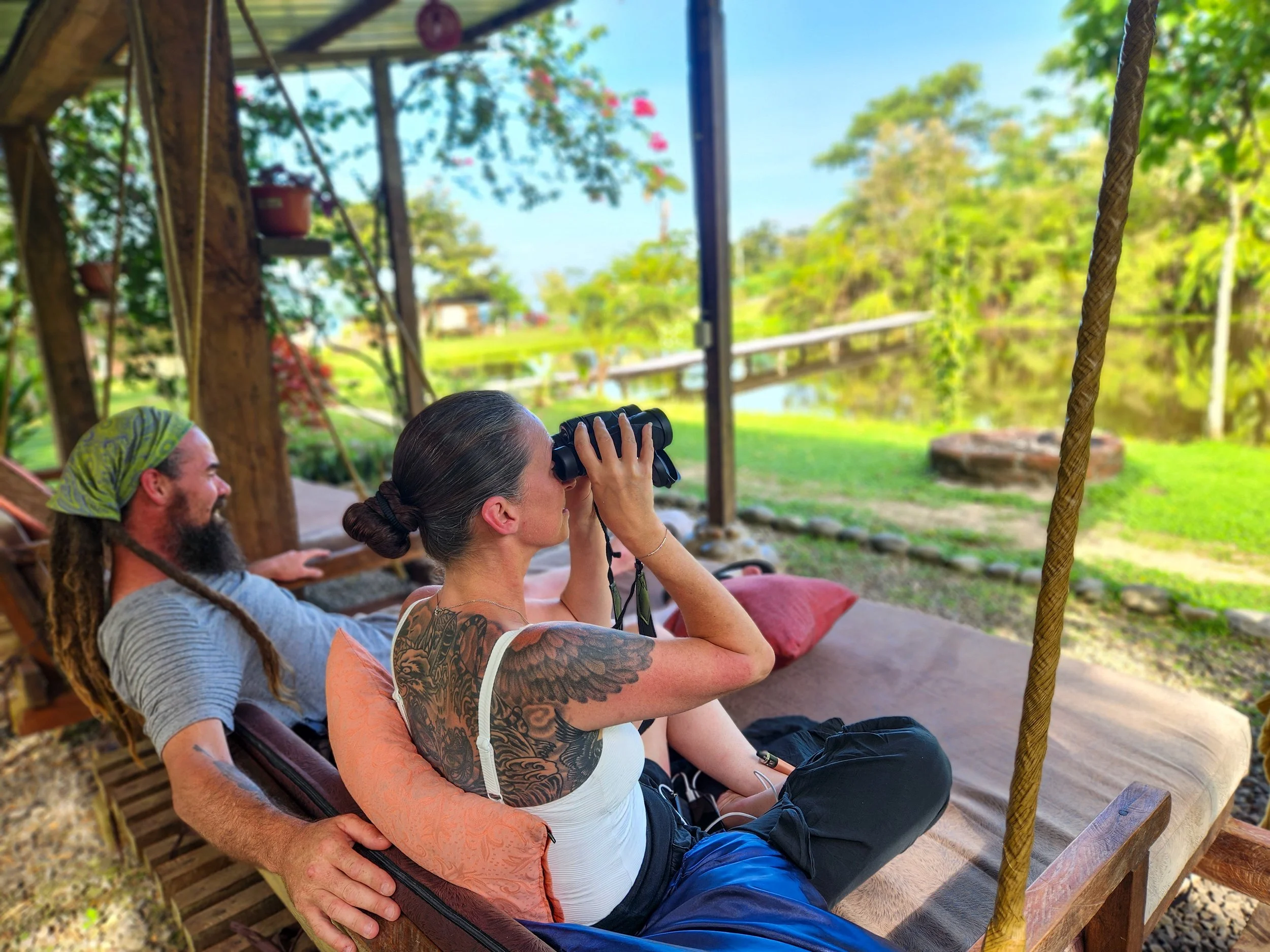 Dos personas sentadas en una cama, una mujer con gafas de binoculares y un tatuaje en el brazo, y un hombre con barba y pañuelo en la cabeza, observando un entorno natural con árboles y agua.
