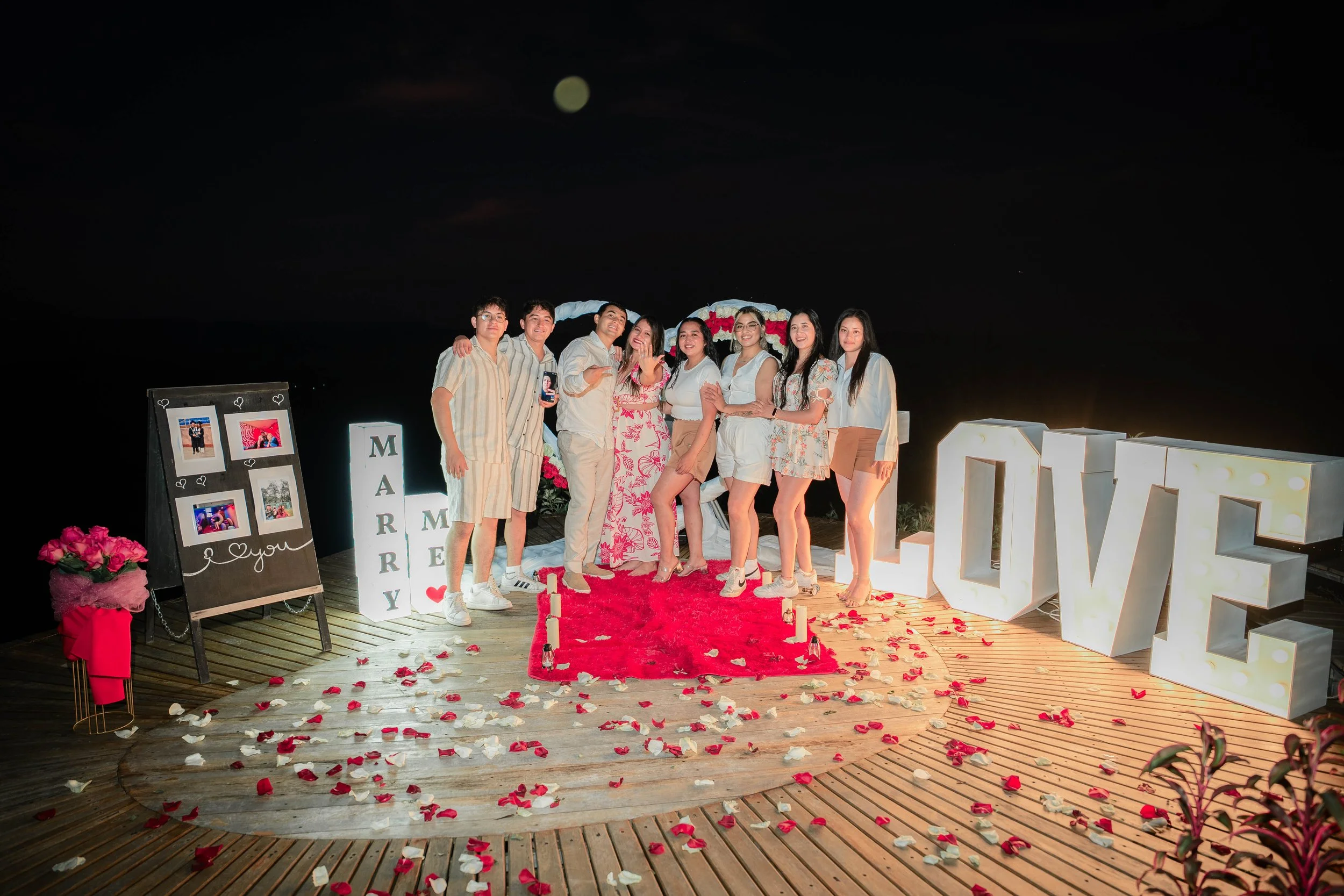 Grupo de personas en una celebración de aniversario, rodeados de decoraciones de corazones, flores y letras grandes que dicen 'LOVE', en una plataforma de madera